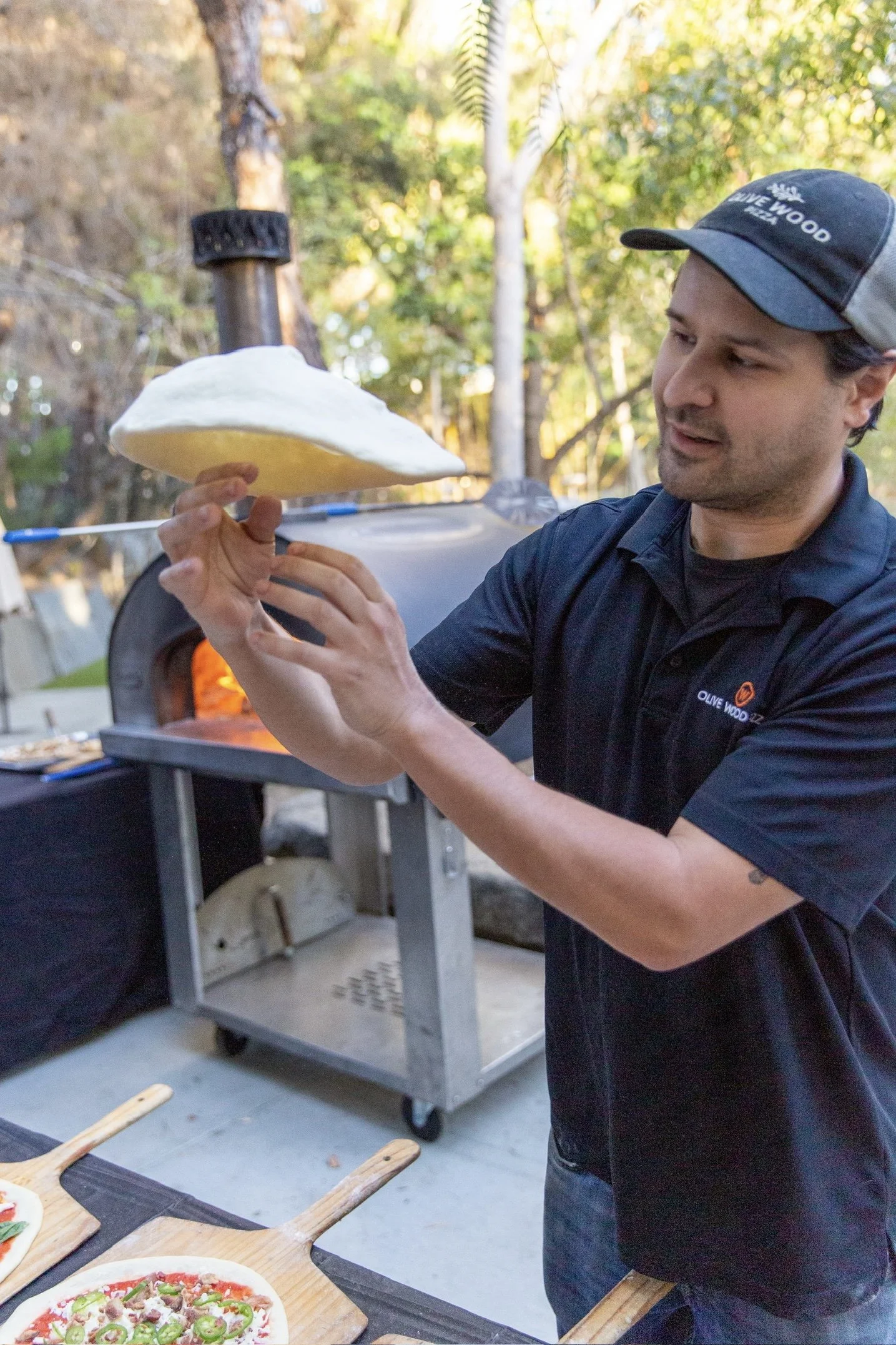 A man stretching a pizza, standing outdoors near a wood-fired pizza oven, with trees and sunlight in the background.