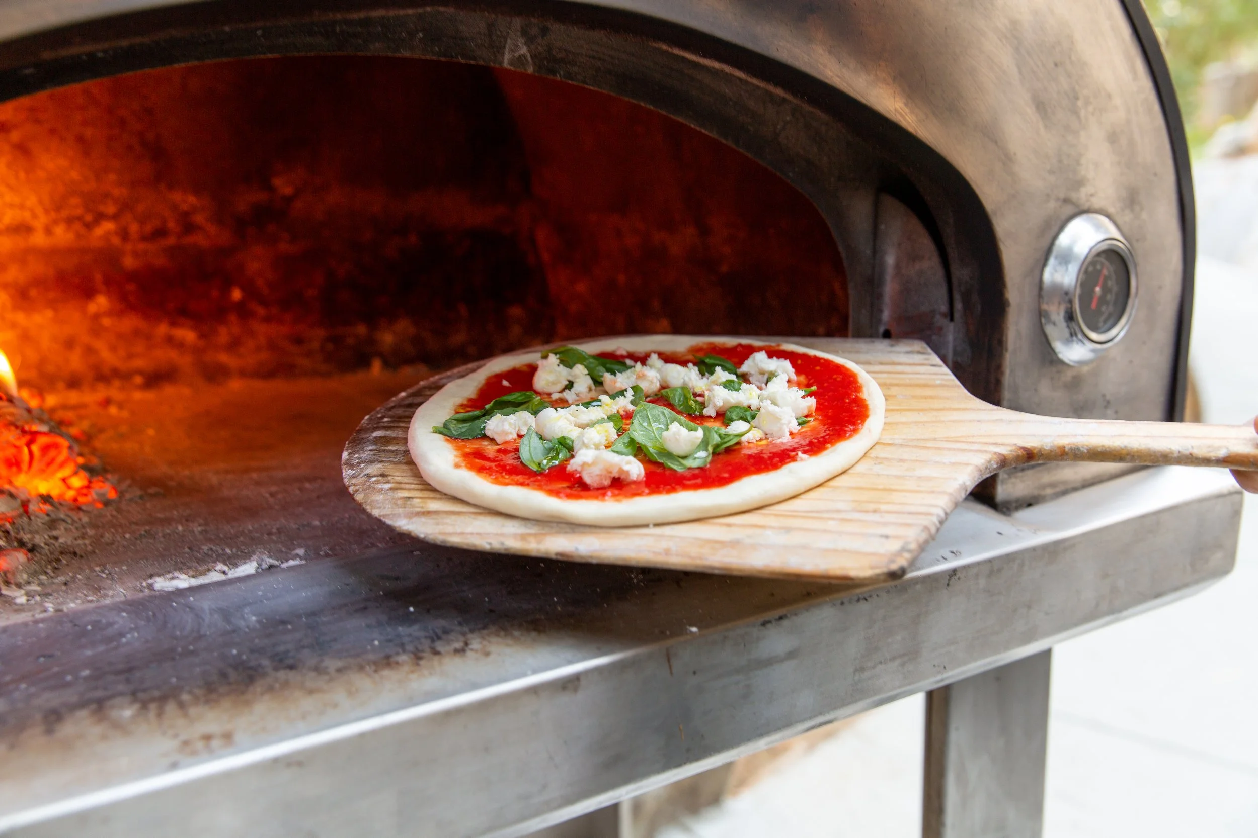 Pizza being placed into a wood-fired oven on a peel during live catering by Olive Wood Pizza