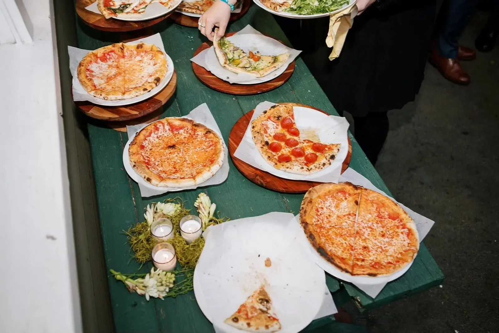 Several pizzas on wooden and paper plates on a green table, some with cheese and crushed tomato, one with pepperoni slices, a hand removing a slice, and some small lit candles with flowers in front.