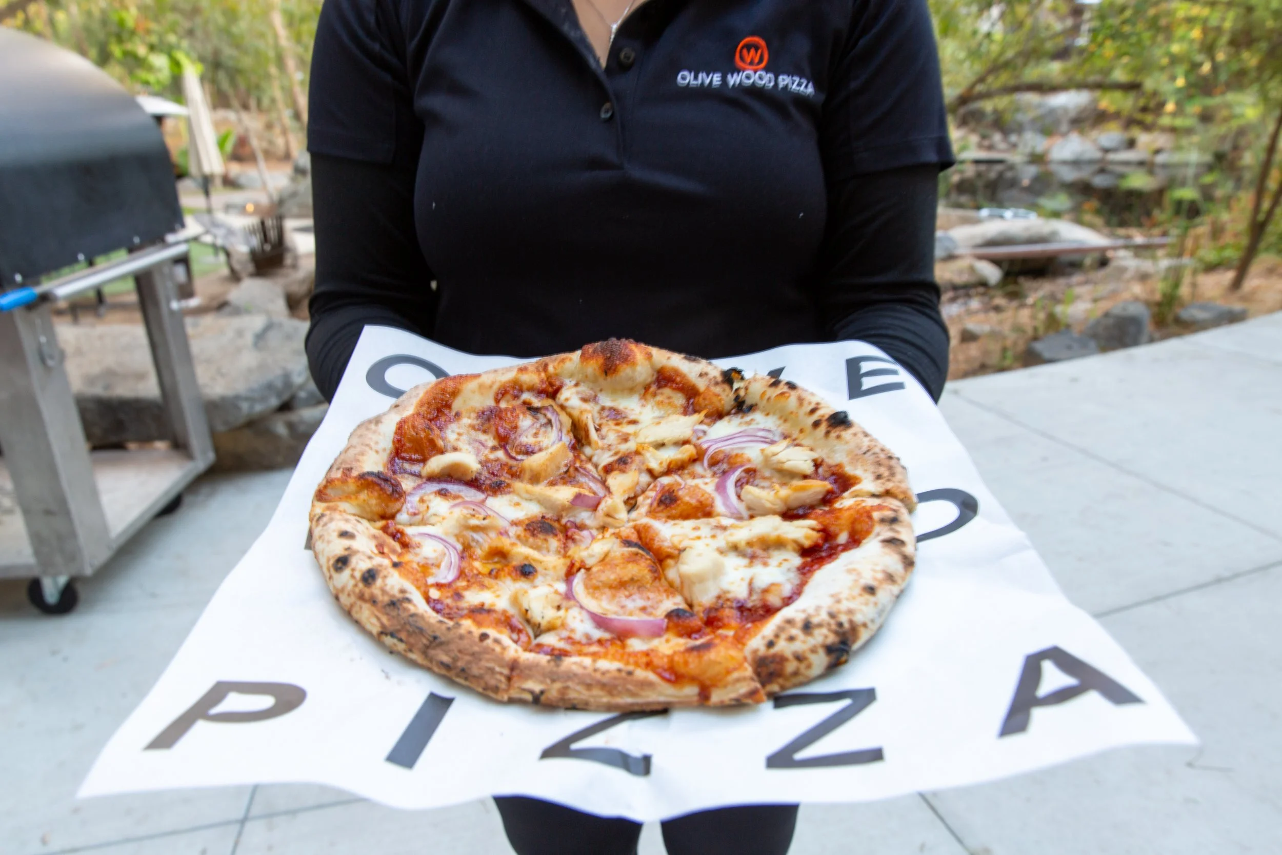Person holding a pizza with a cheese, chicken, red onion and cilantro pizza, outdoors, wearing a black polo shirt with an "Olive Wood Pizza" logo.