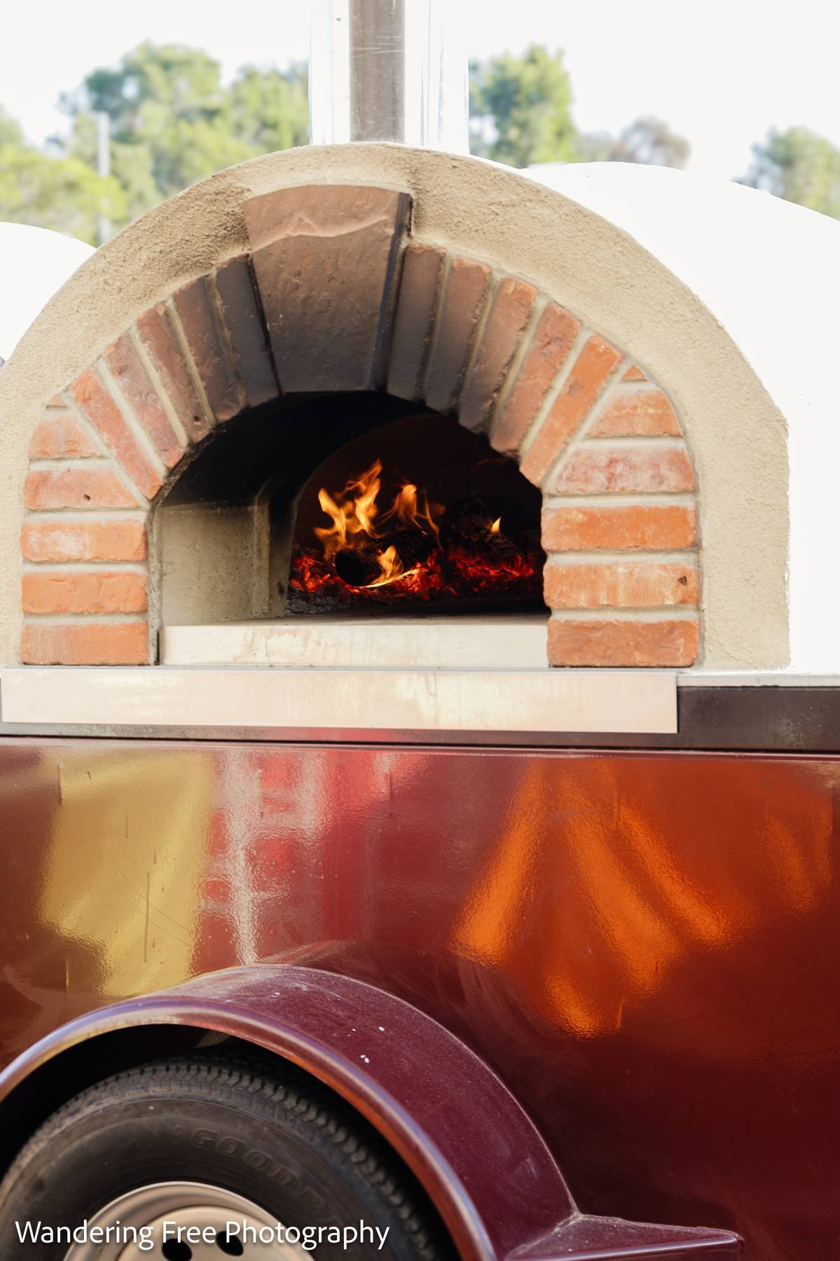 A brick and stone outdoor pizza oven with a fire burning inside, mounted on top of trailer with a tire visible at the bottom.