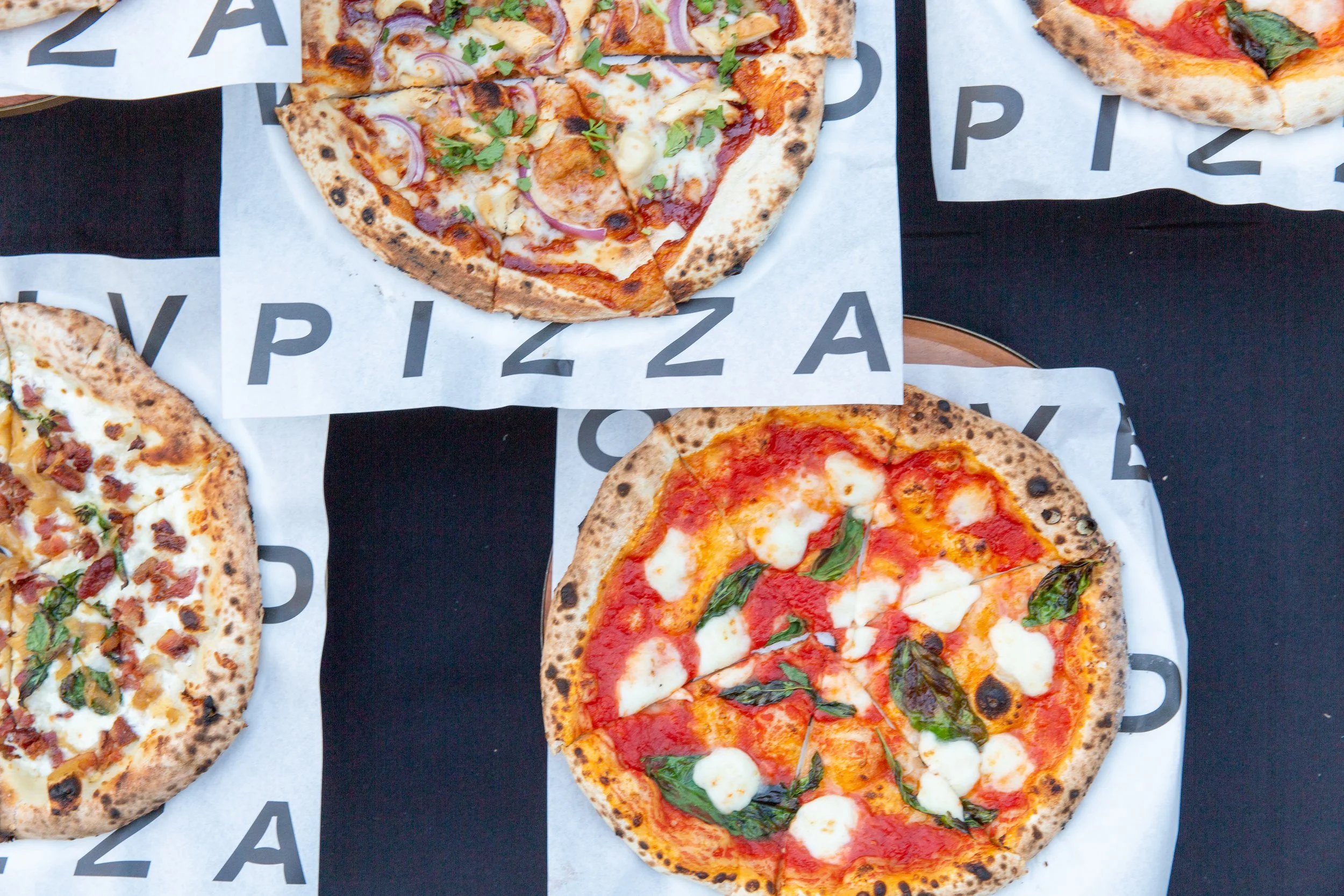 Assorted wood-fired pizzas displayed on branded paper at an Olive Wood Pizza event