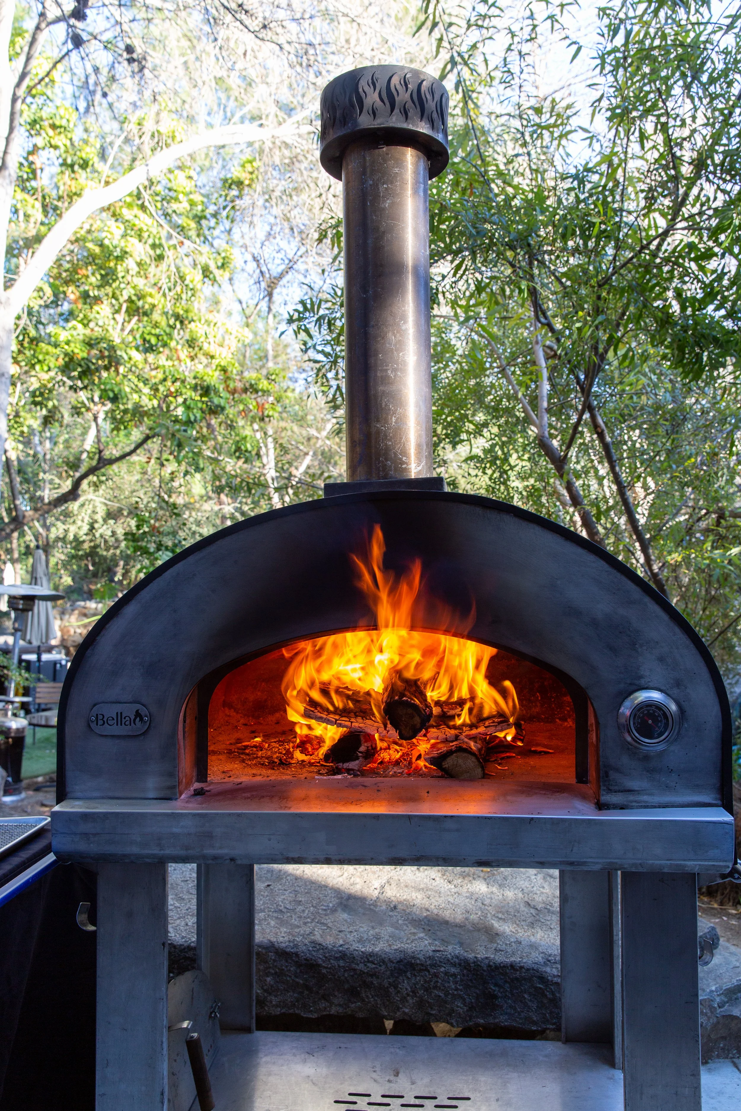 A wood-fired pizza oven with flames inside, made of stainless steel with a smoke chimney on top, situated outdoors among trees.