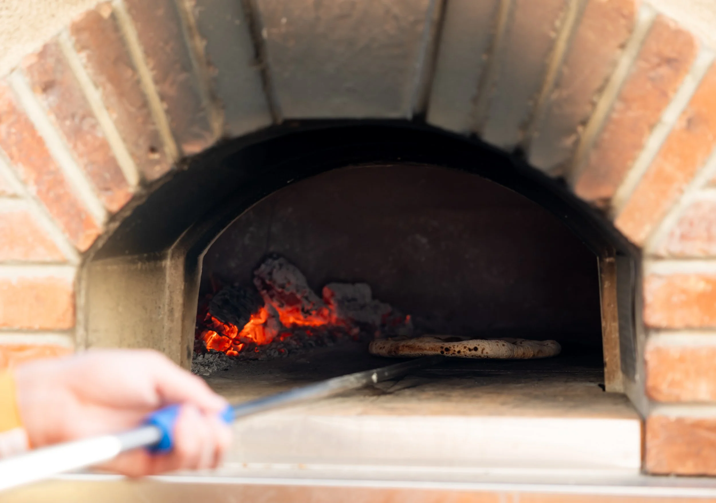 Pizza being placed into a wood-fired brick oven using a peel by Olive Wood Pizza