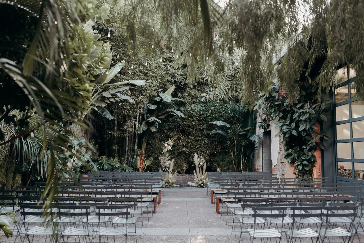 Outdoor Marvimon venue space with rows of empty chairs facing a floral arch, surrounded by lush green plants and hanging string lights.