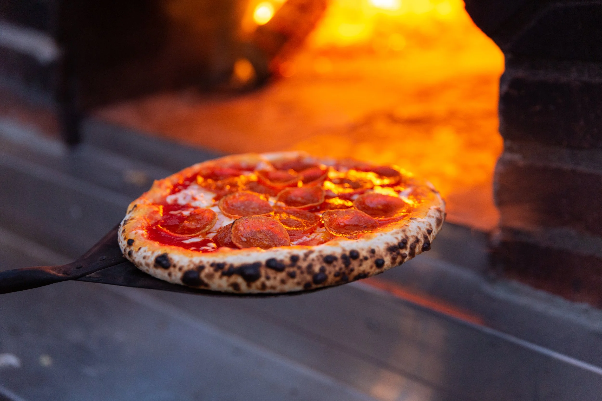 Pepperoni pizza cooking in a wood-fired oven at an Olive Wood Pizza catering event
