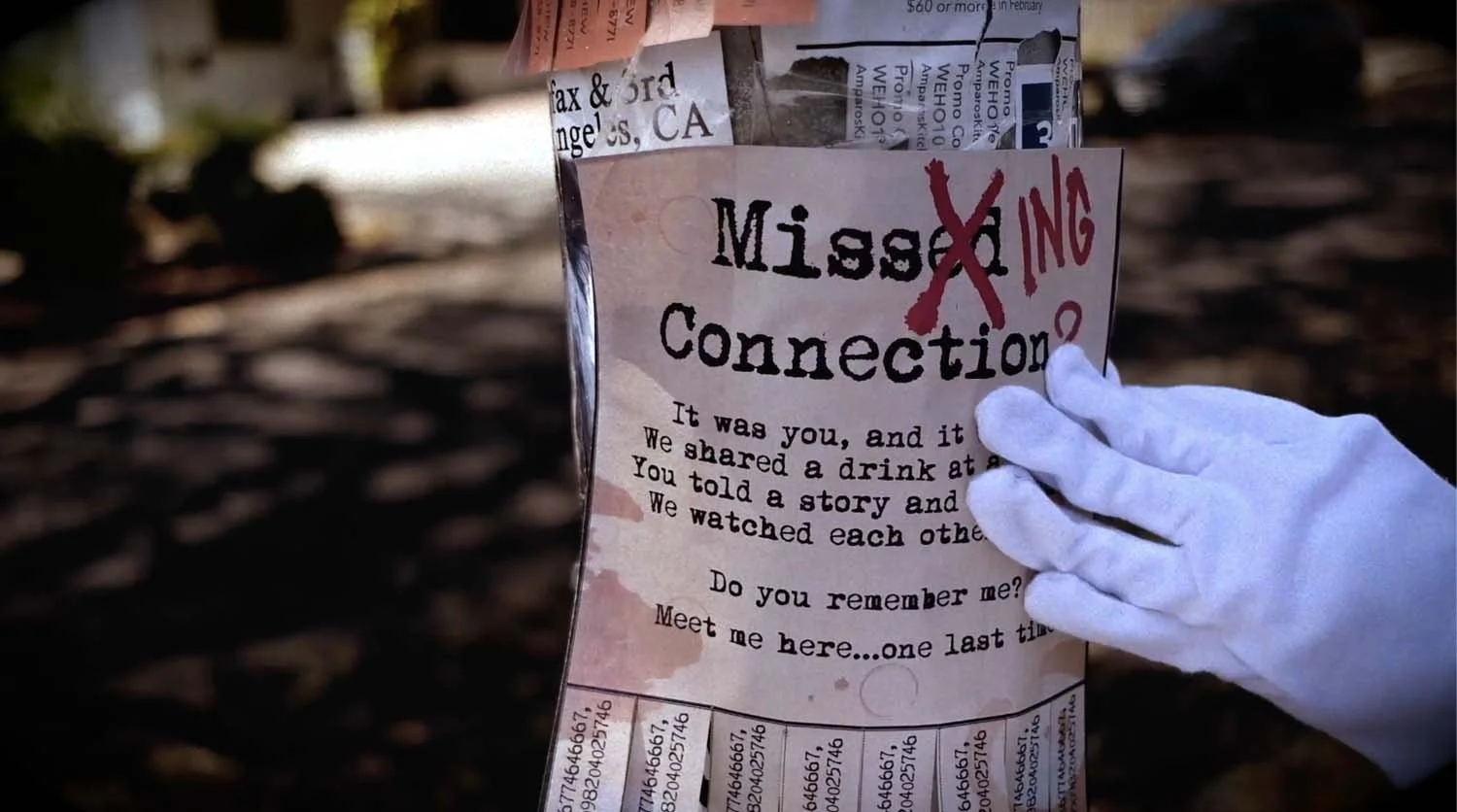 A person's hand in a white glove holding a torn paper with a message about shared experiences and a reconnecting invitation. The paper has the words 'Missing Connection' with the 'X' crossed out in red, and is outdoors with a blurred background of soil and trees.