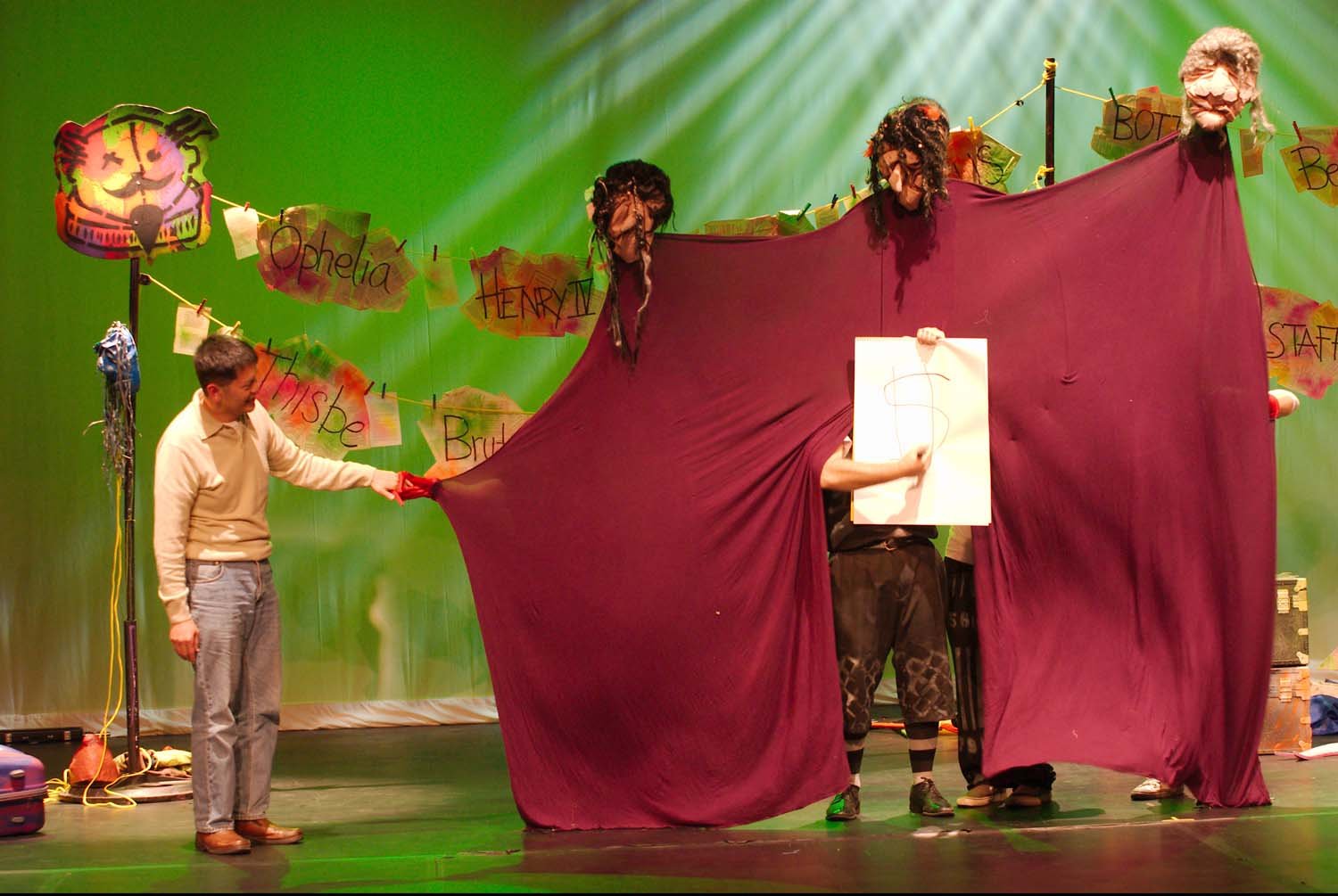 The scene depicts a stage with a green backdrop. Several people are involved in what appears to be a puppetry performance. A large curtain is being manipulated by individuals behind it, with one person holding a whiteboard with the letter 'S' written on it. The backdrop is decorated with colorful signs and heads of puppets with various hairstyles and facial features. One person on the left is holding part of the curtain, smiling, and the overall scene suggests a playful or theatrical presentation.