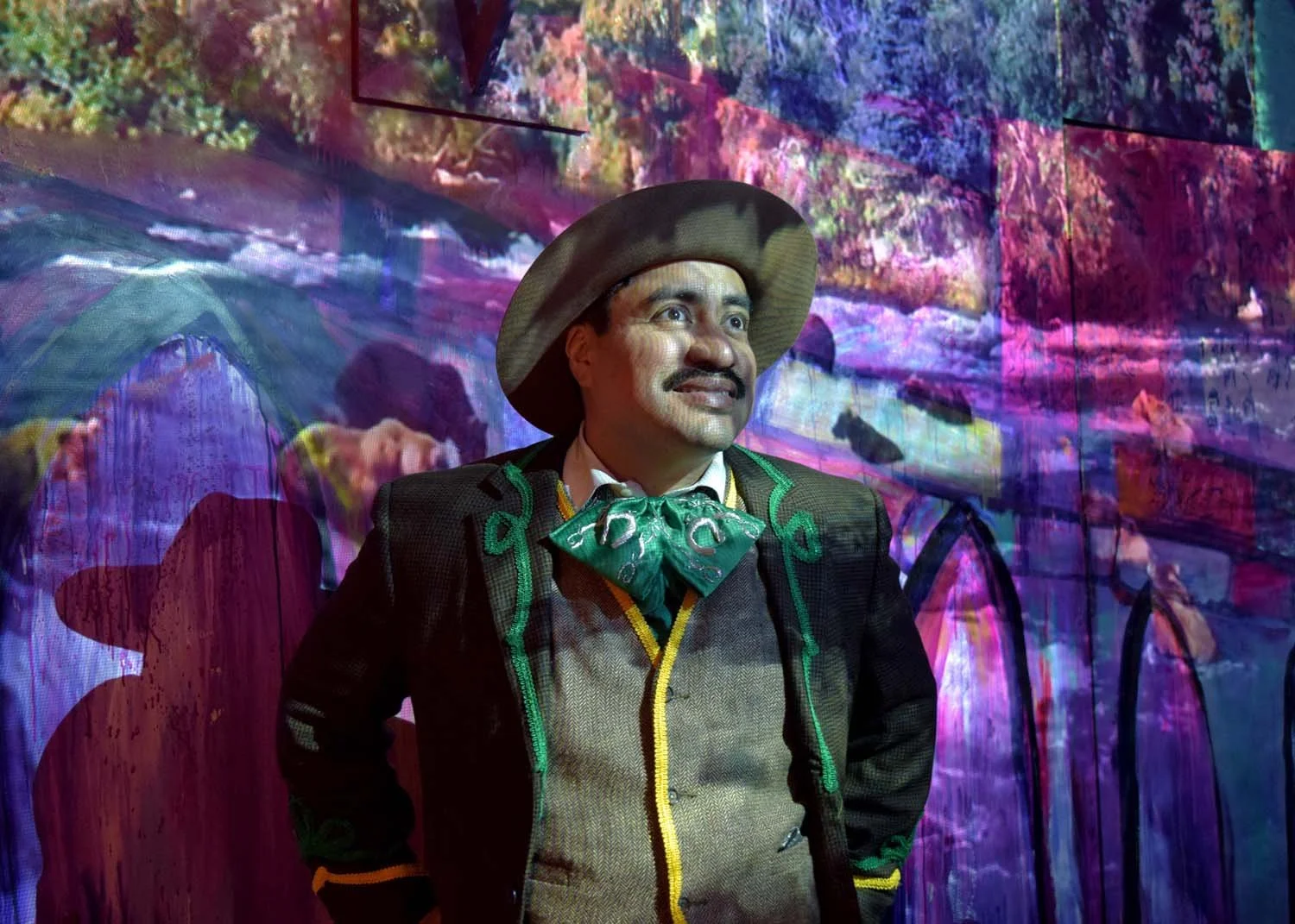Man dressed as a Mexican charro, wearing a hat, a jacket with green embroidery, and a green bow tie, standing in front of a colorful, digitally projected background with abstract shapes and vibrant colors.