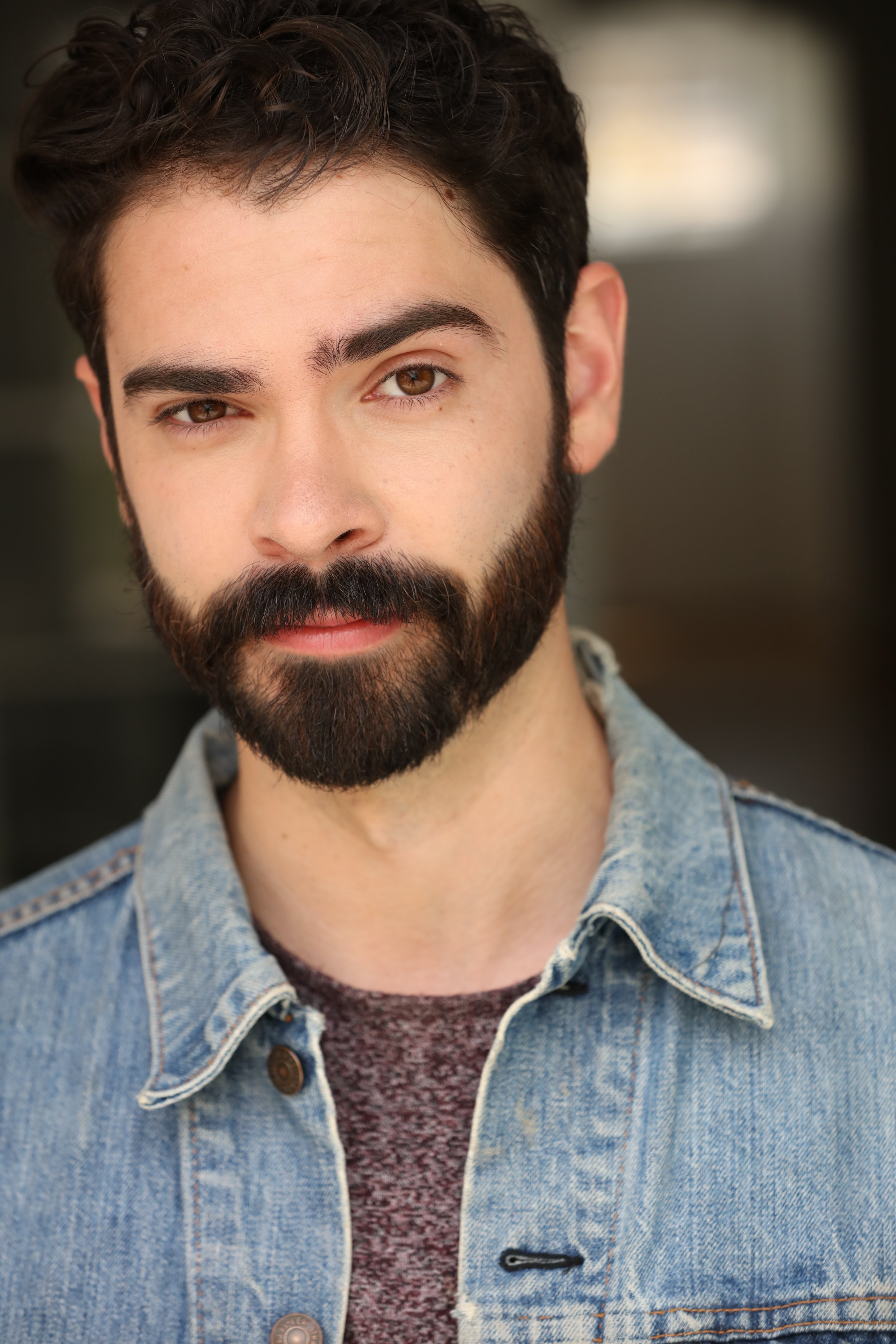 Close-up of a young man with dark curly hair, brown eyes, and a beard, wearing a light blue denim jacket over a maroon sweater, in an indoor setting with blurred background.