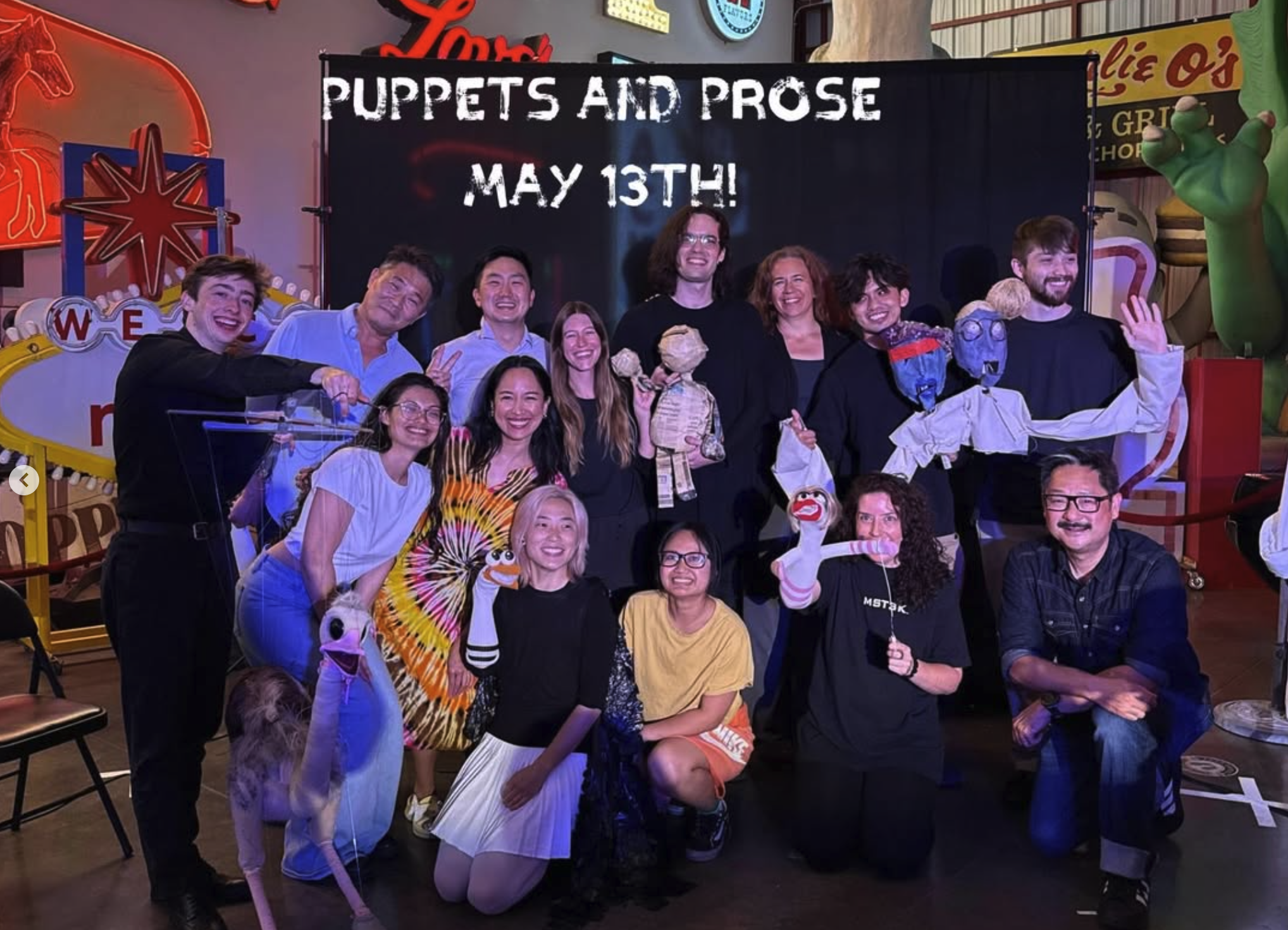 Group of people posing in front of a black banner with white text that reads 'Puppets and Prose May 13th!'. The group includes individuals holding puppets and smiling. The background features neon signs and colorful decorations.