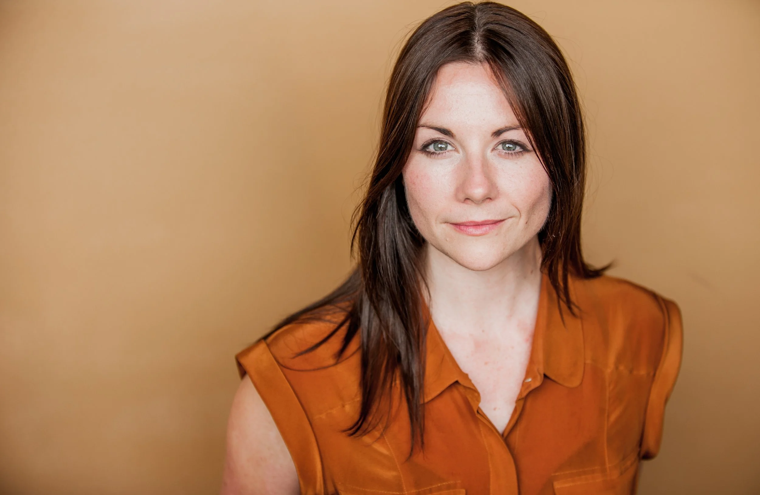 A woman with long dark hair and blue eyes smiling, wearing an orange top, against a plain beige background.