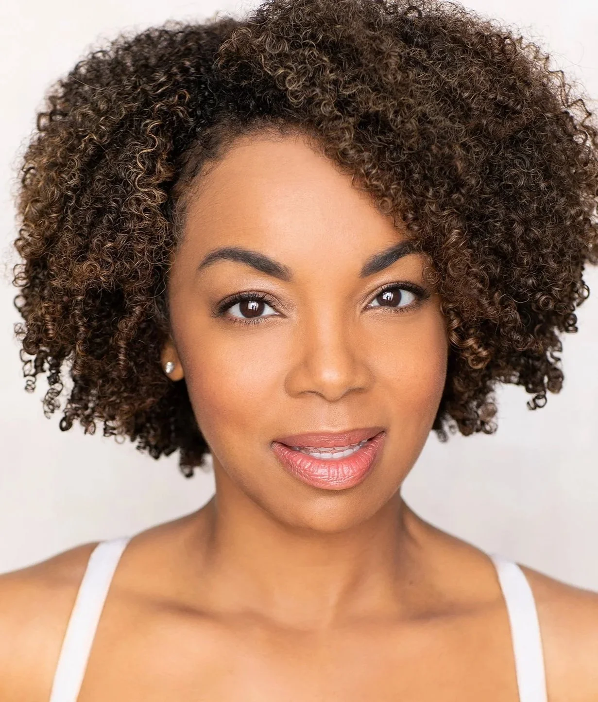 Close-up portrait of a woman with curly brown hair, brown eyes, and wearing pearl earrings and a white spaghetti strap top, smiling softly against a light background.