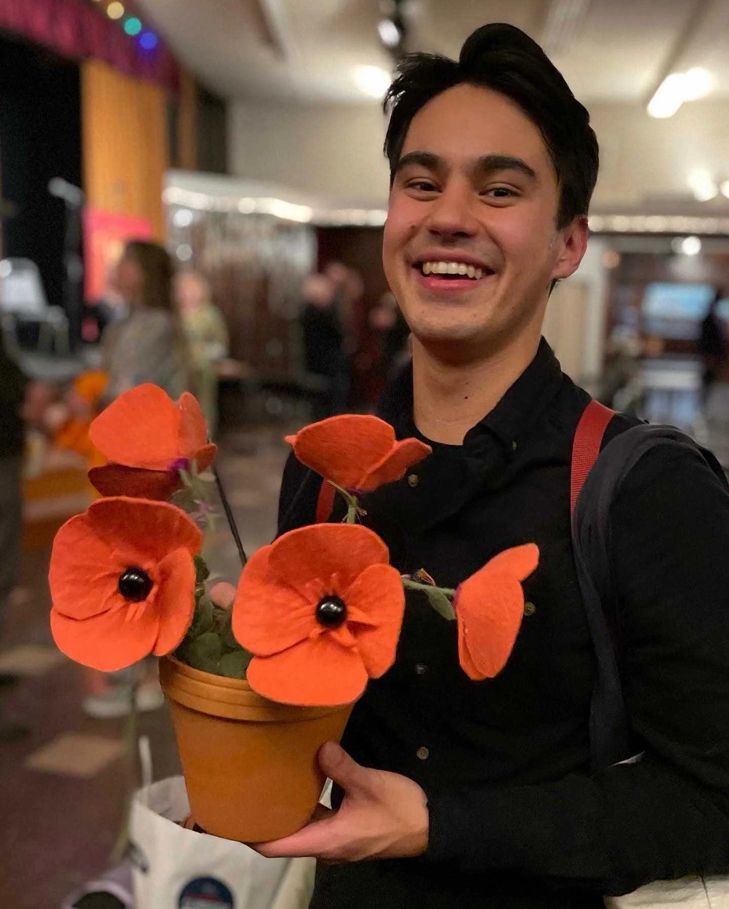 A young man smiling while holding a pot of orange flowers with black centers in a dimly lit indoor setting.