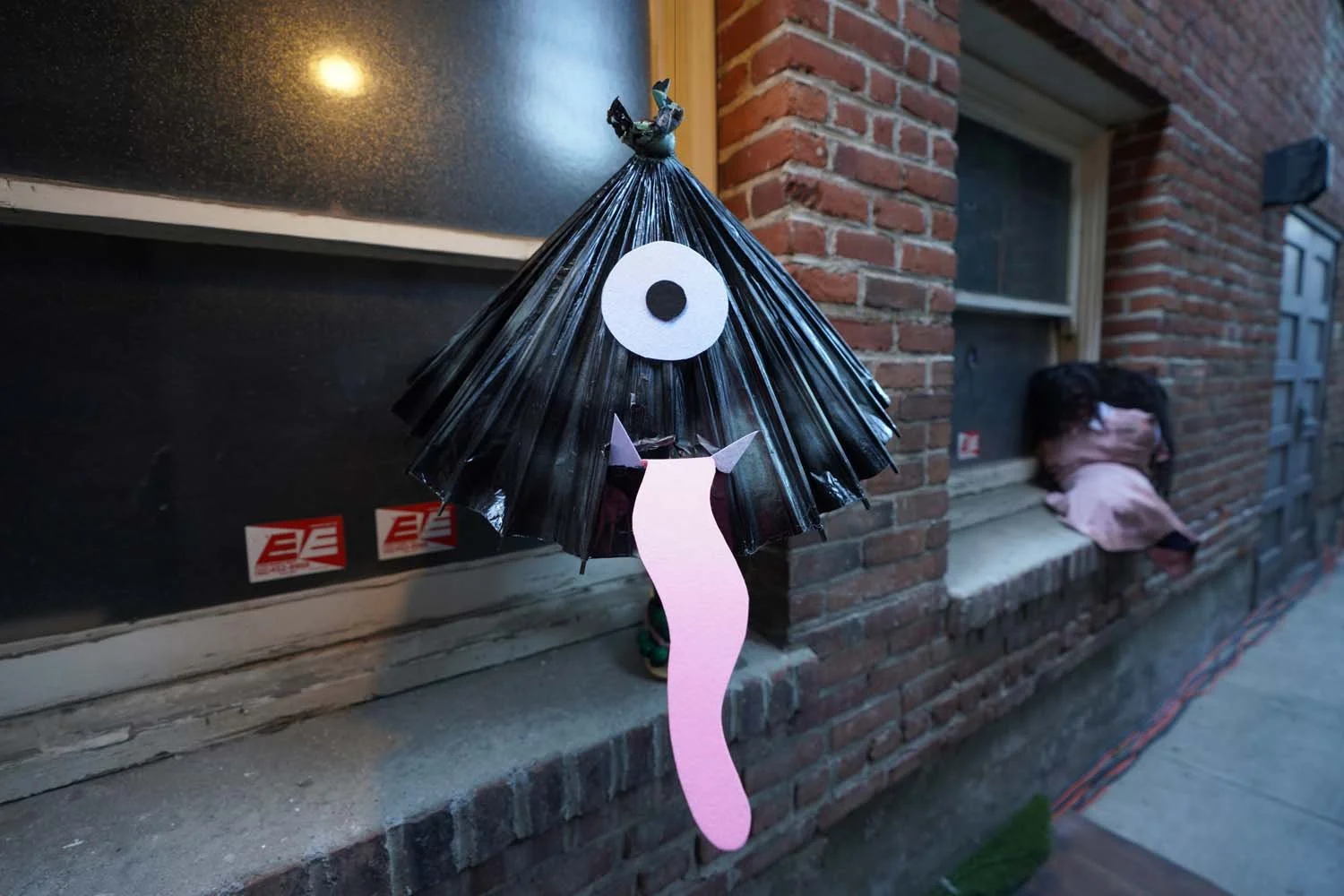 A handmade Halloween decoration of a cat with a black paper umbrella body, a white paper eye, a pink paper tail, and black felt ears, hanging outside a brick house, with a person in pink pajamas sitting on the windowsill in the background.