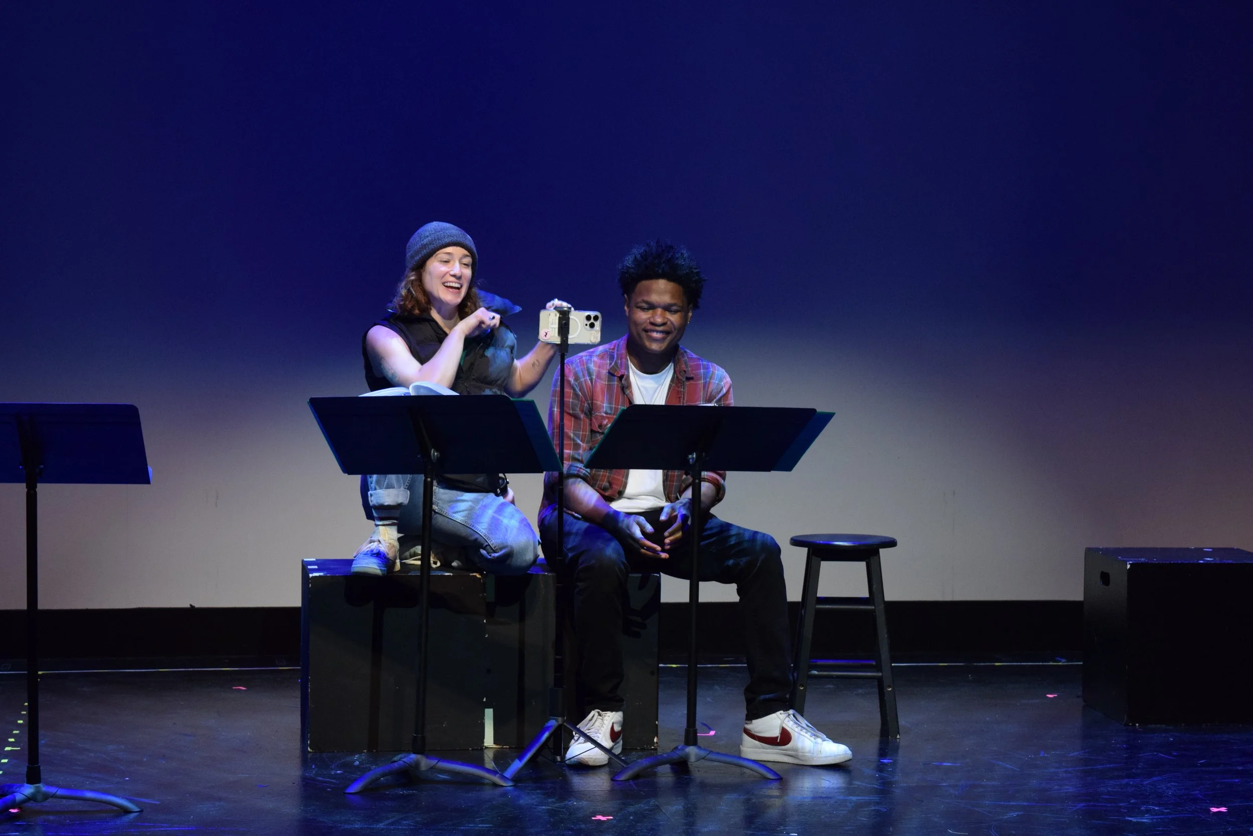 Two people sitting on a stage; the woman is kneeling, holding a smartphone on a stand, and the man is sitting on a stool with a music stand in front of him. The woman is smiling while looking at the phone, and the man is looking down. The background 