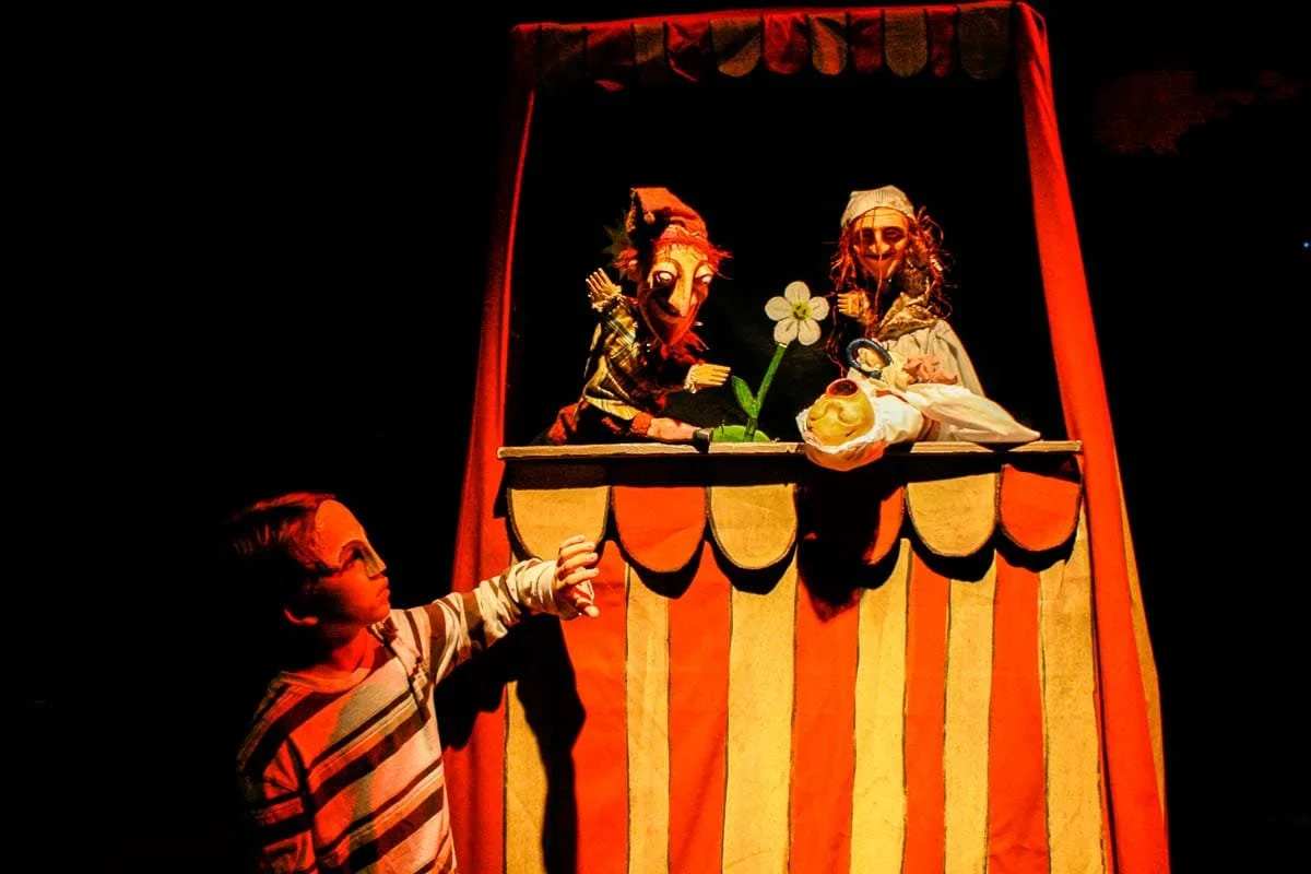 A puppet show with three puppets on a stage decorated with orange and yellow scalloped trim; one puppet is a woman with a bonnet, another puppet is a woman with a crown of leaves, and the third is a woman holding a flower; a young boy is pointing at the puppets from below.