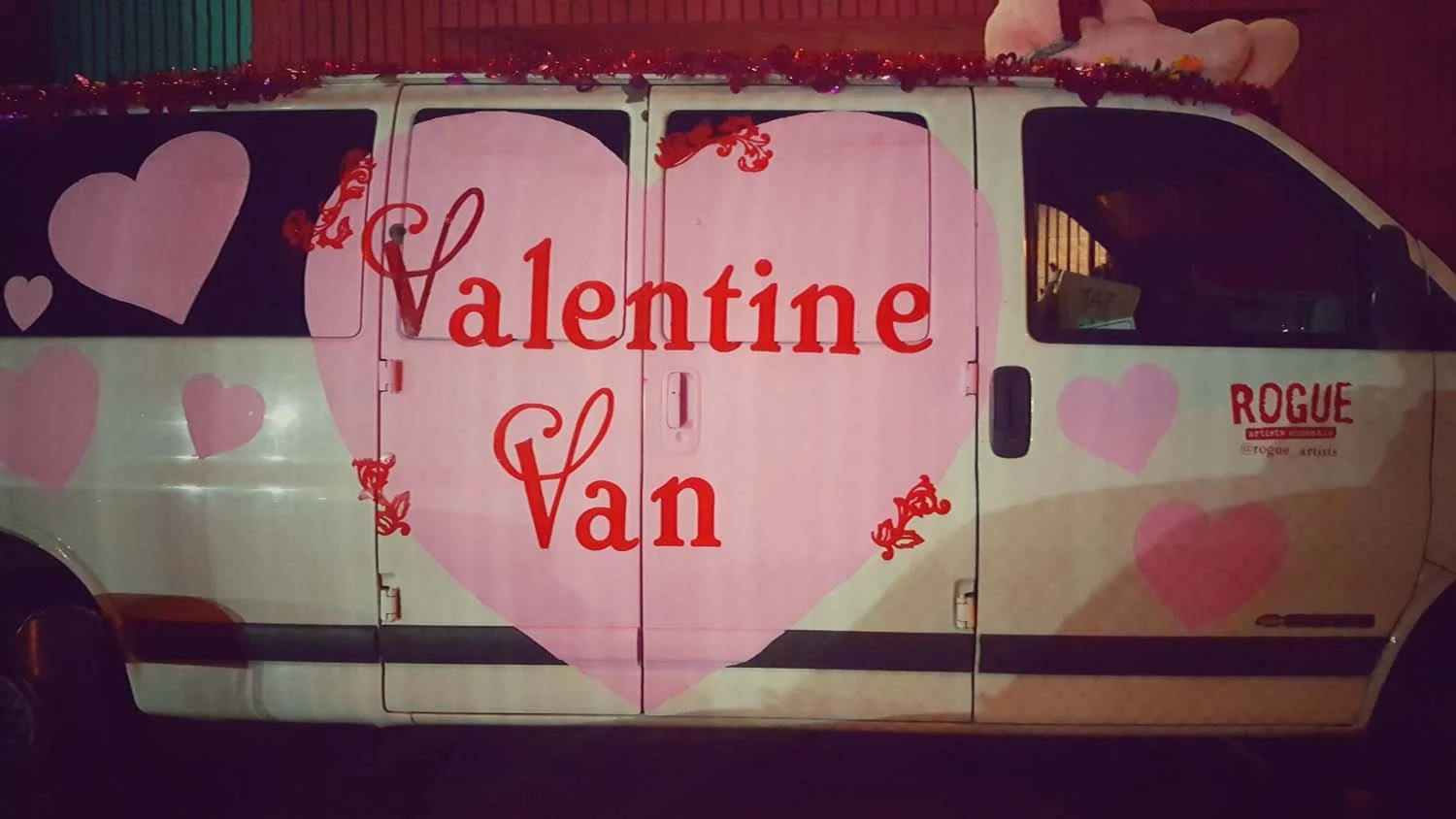 A white van decorated for Valentine's Day with pink and red hearts, and large red text reading 'Valentine Van' in a pink heart. The van also features the name 'Rogue' and social media handle '@rogue_artists' on the front side.