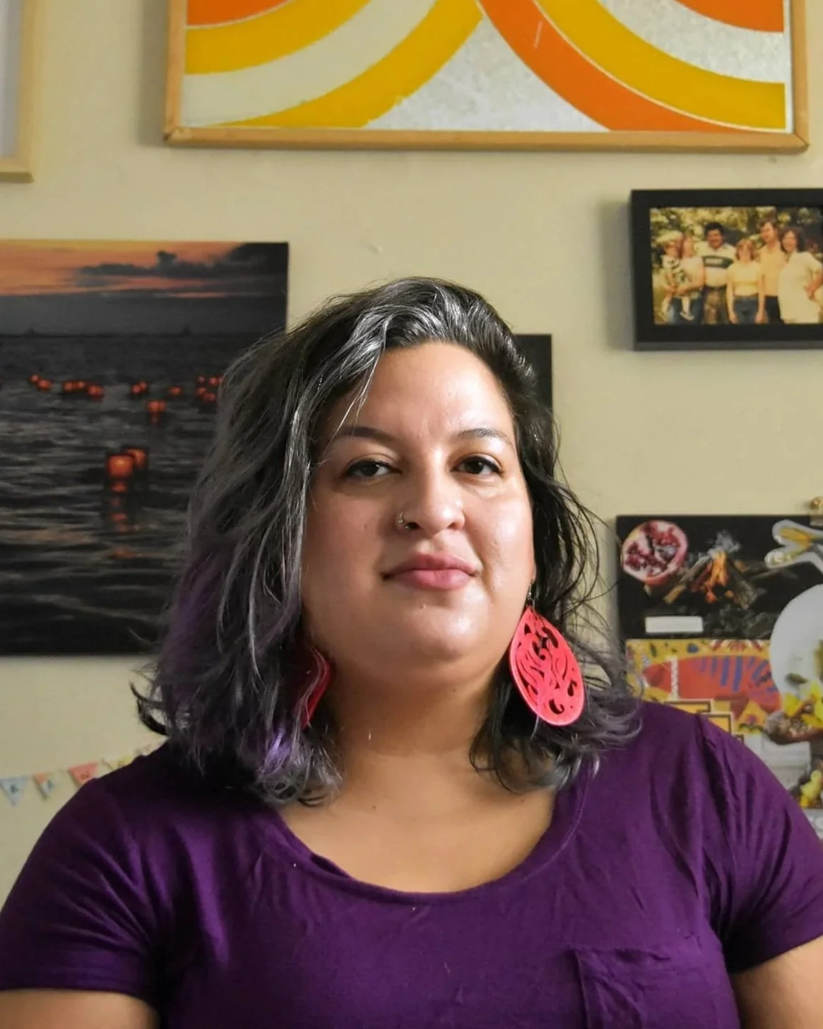 A woman with shoulder-length dark hair with purple streaks, wearing a purple top and large red earrings, standing in front of a wall with framed photographs and artwork.