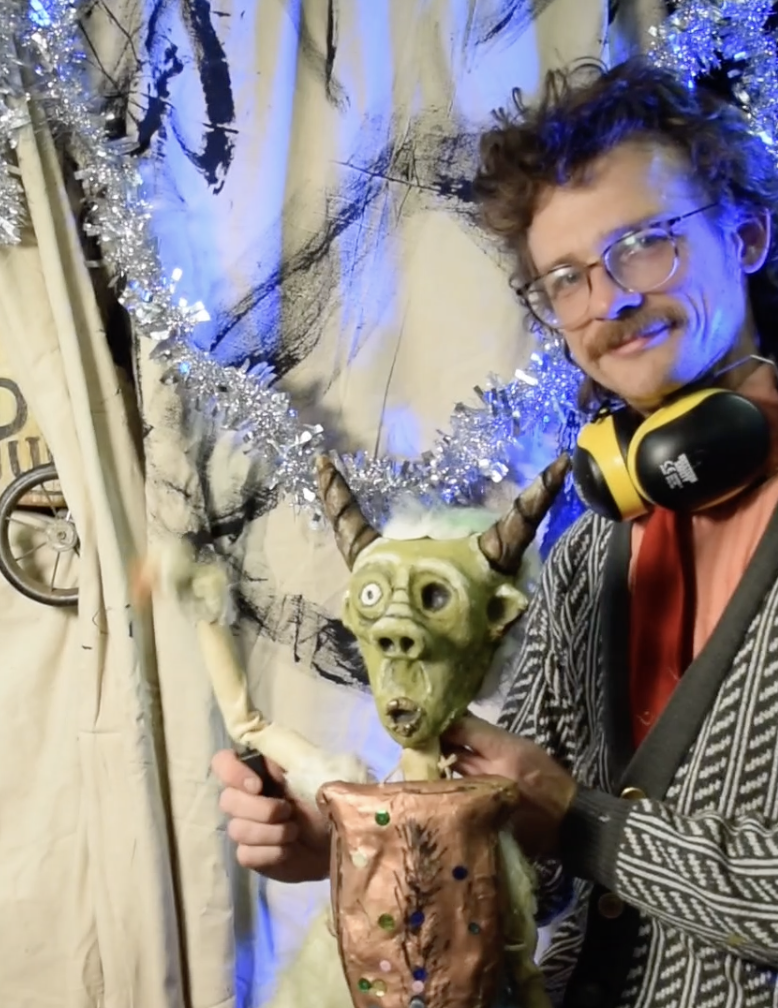 A man with glasses and curly hair wearing headphones, holding a puppet of a goblin-like creature with green skin, large eyes, and horns, in front of a whimsical backdrop with a tinsel decoration.