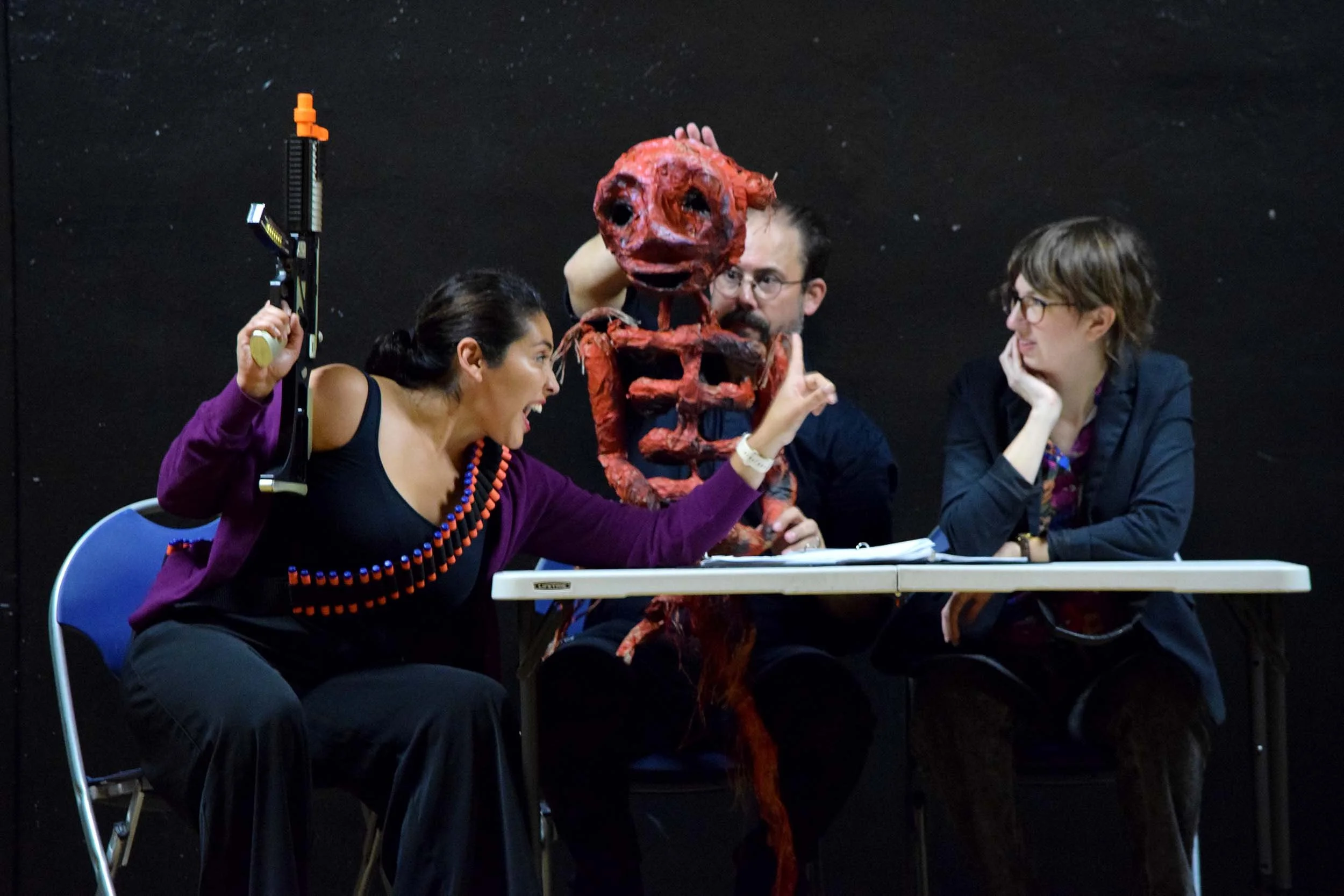 Three people sitting at a table with a large, red, grotesque puppet or mask of a creature with an open mouth and big eyes. They appear to be in discussion or performing a scene, with one woman holding a prop or weapon and another speaking, while the third person looks on. The backdrop is black with stars.