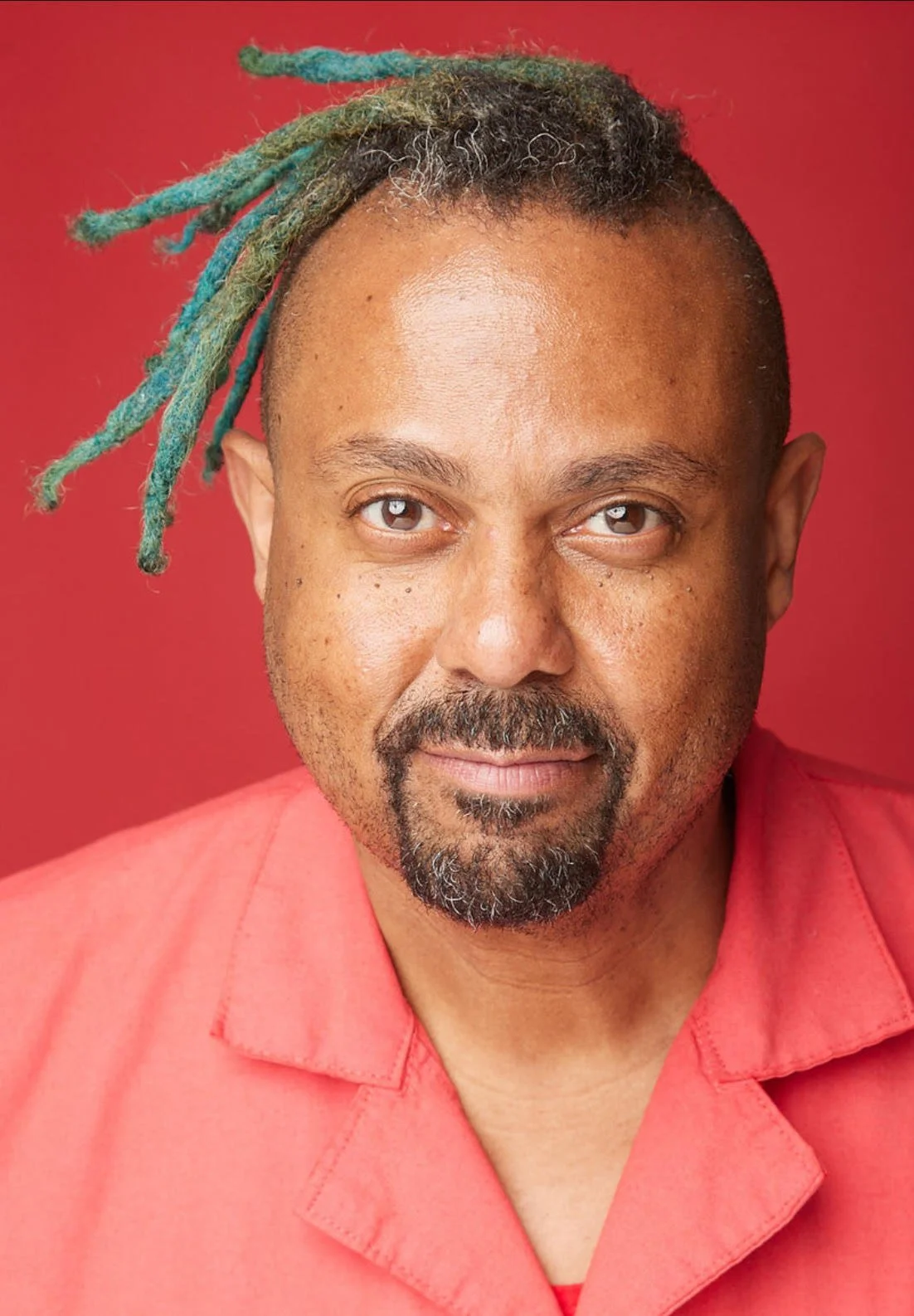 A man with medium brown skin, short gray beard, and dreadlocked hair with some colored blue and green dreadlocks, wearing a coral-colored shirt, looking directly at the camera against a red background.
