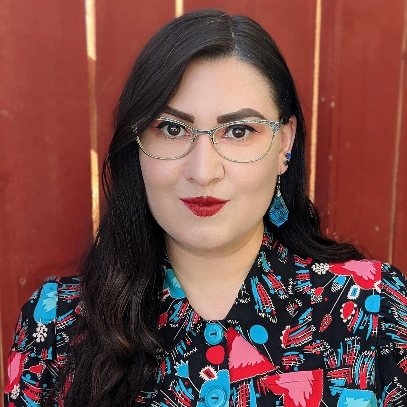 A woman with long dark hair, wearing glasses, red lipstick, and a colorful patterned blouse, standing in front of a wooden background.