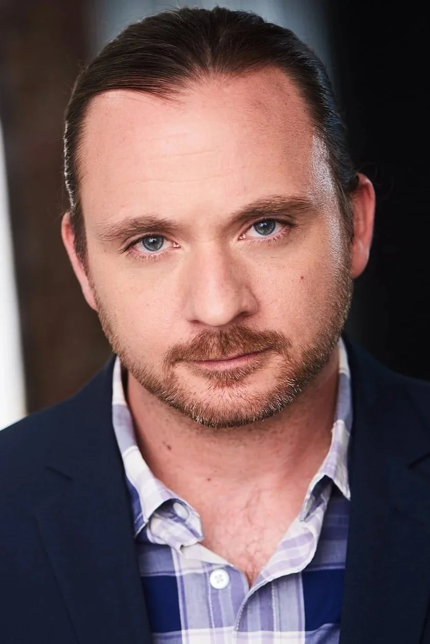 Close-up portrait of a man with light skin, blue eyes, dark brown hair slicked back, a beard, wearing a dark blazer and a light blue plaid shirt, in an indoor setting.