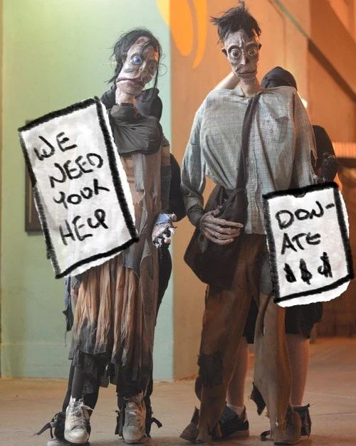 Two mannequins dressed as homeless people with signs. The left sign reads 'We need your help', and the right sign reads 'Don't eat' with dollar signs.