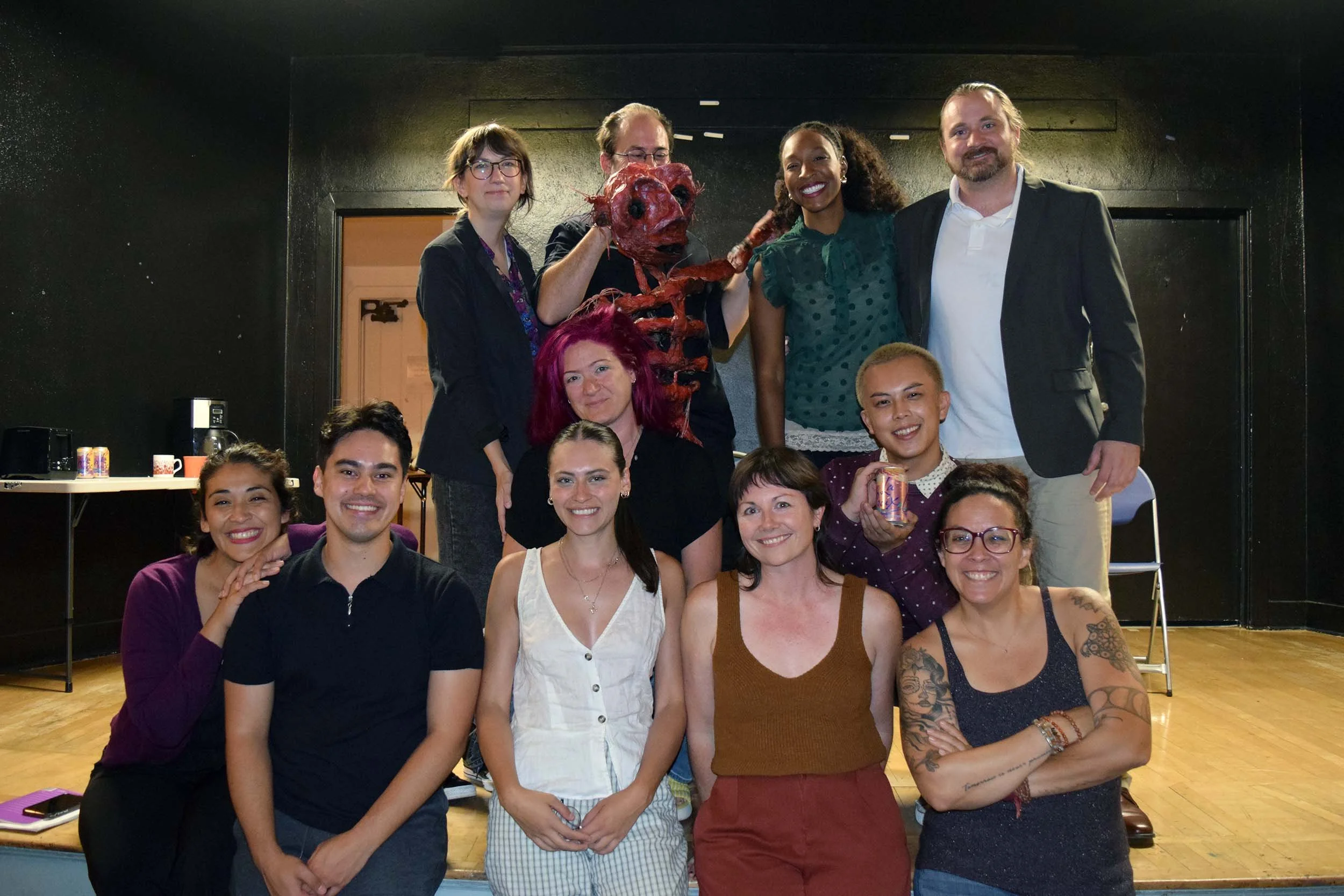 A group of diverse people posing and smiling in a room, with some holding drinks, and a person in the back wearing a costume resembling a creature with a red, pig-like face and a long, humanoid body.