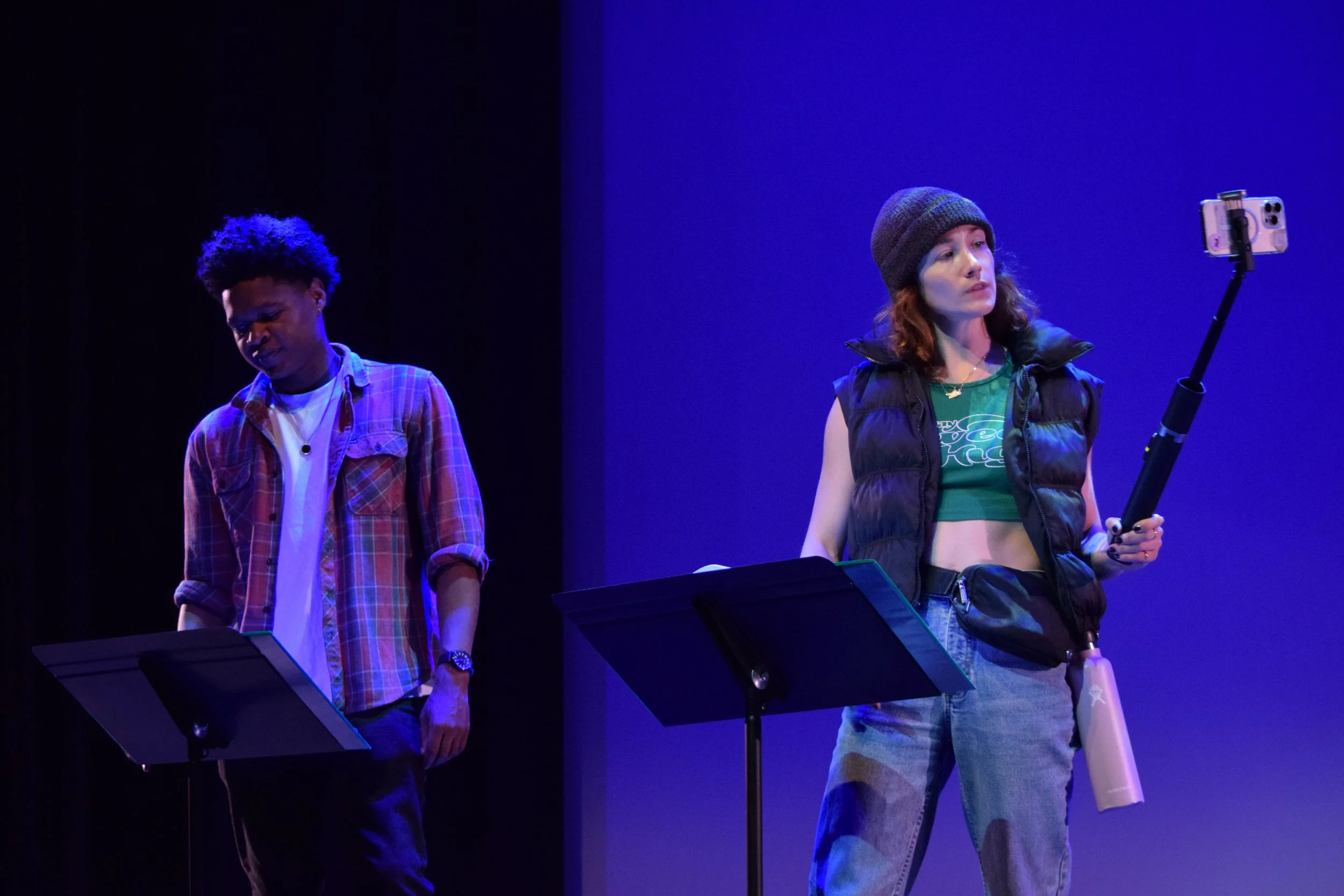 Two women performing on stage with music stands, one wearing a plaid shirt, and the other wearing a beanie and a vest, using a smartphone on a selfie stick.