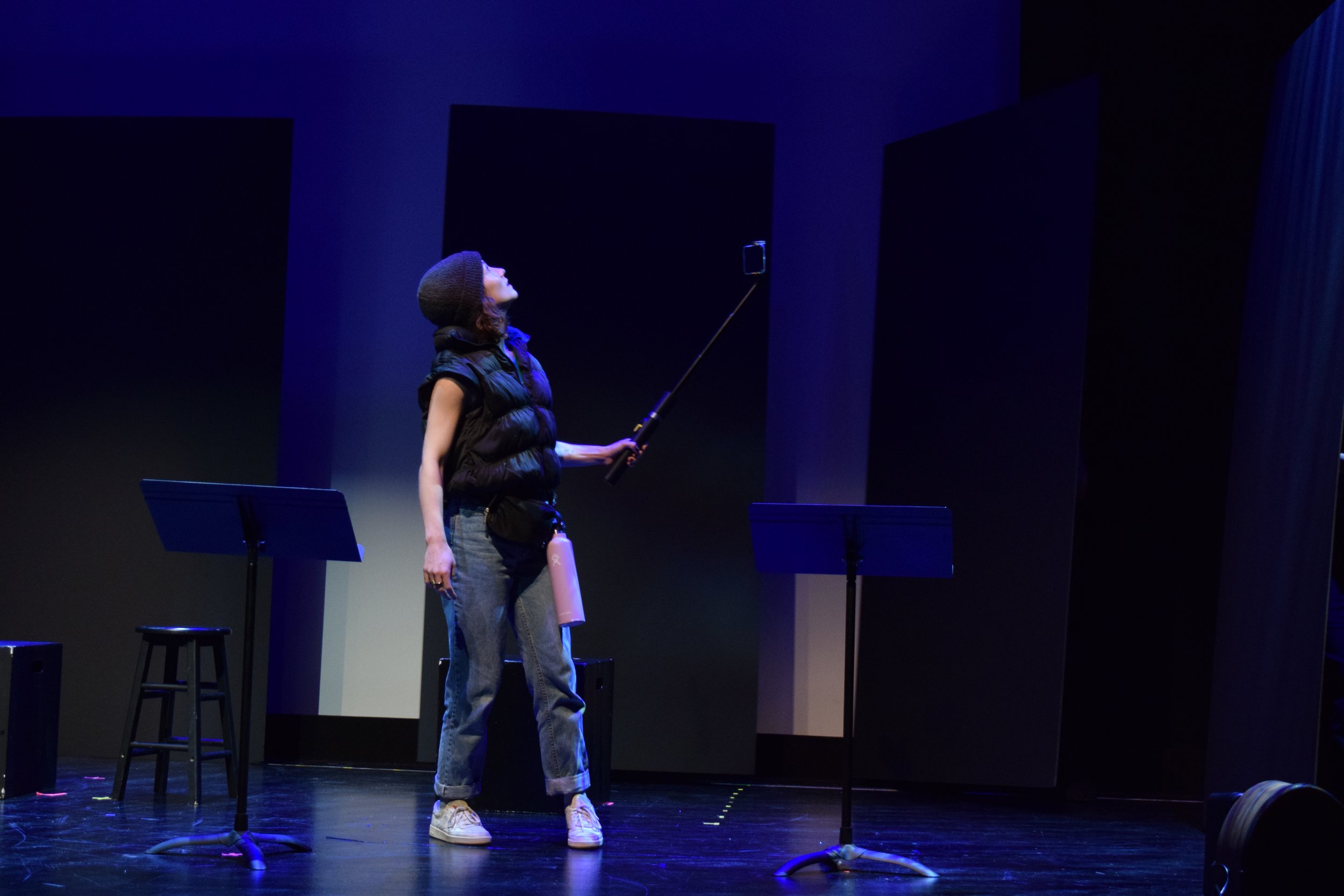 A woman on stage wearing a hat, puffy vest, and jeans, holding a selfie stick with a phone, with chairs and music stands around her, under blue lighting.