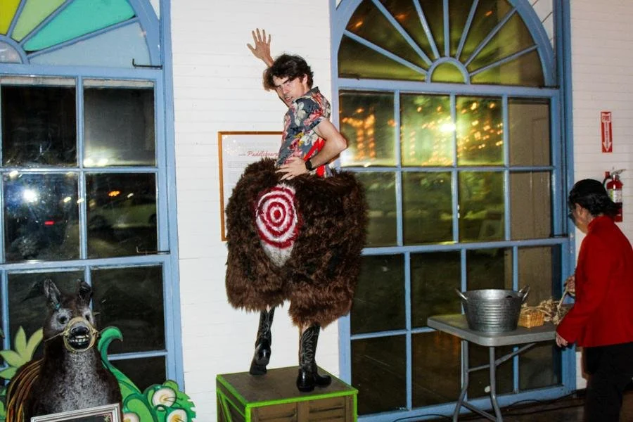 A man standing on a small platform, dressed in a fur costume with a target design on the back, is waving and leaning on a woman dressed in red. The woman is near a table with a metallic bucket, and a decorative horse figure is in the background inside a building with large windows.