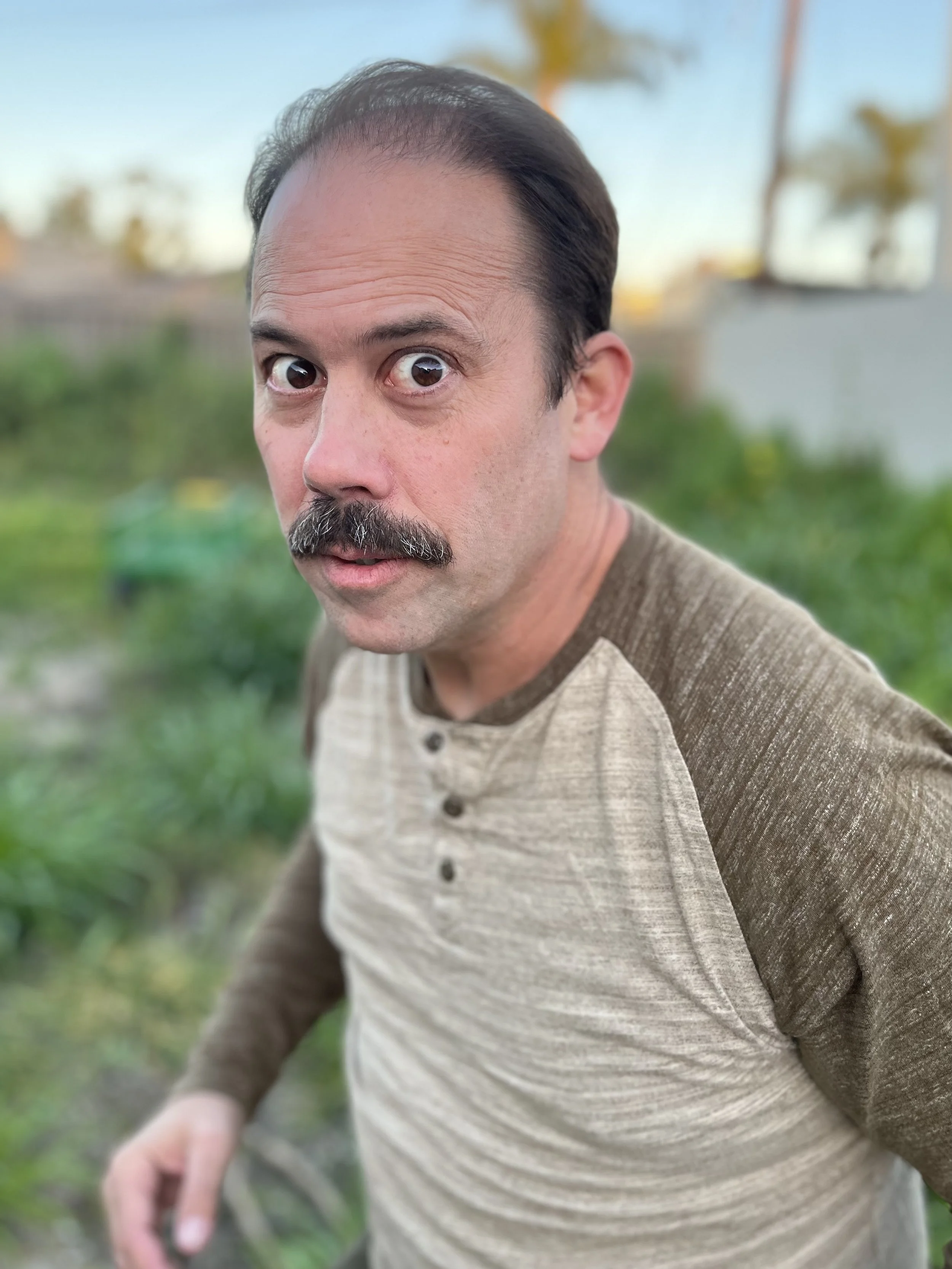A man with a mustache and short dark hair, wearing a beige and brown long-sleeve shirt, looking directly at the camera with a surprised expression, outdoors on a sunny day with blurred greenery and palm trees in the background.