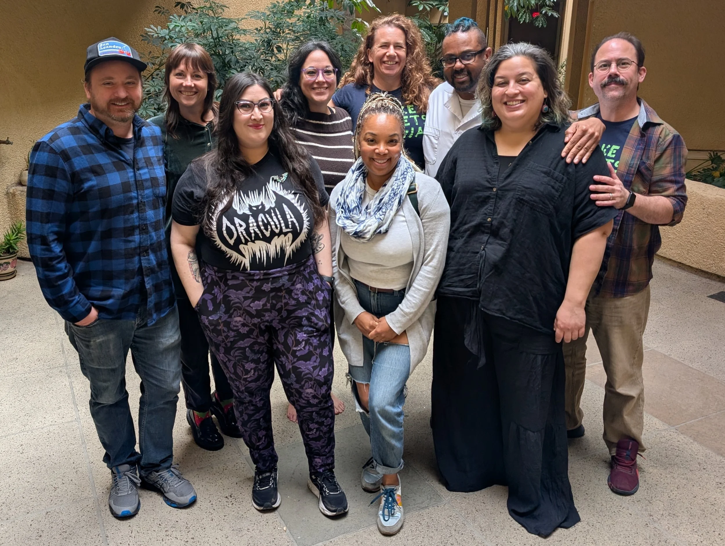 Group of ten diverse people posing together outside with plants and a yellow wall in the background, smiling at the camera.