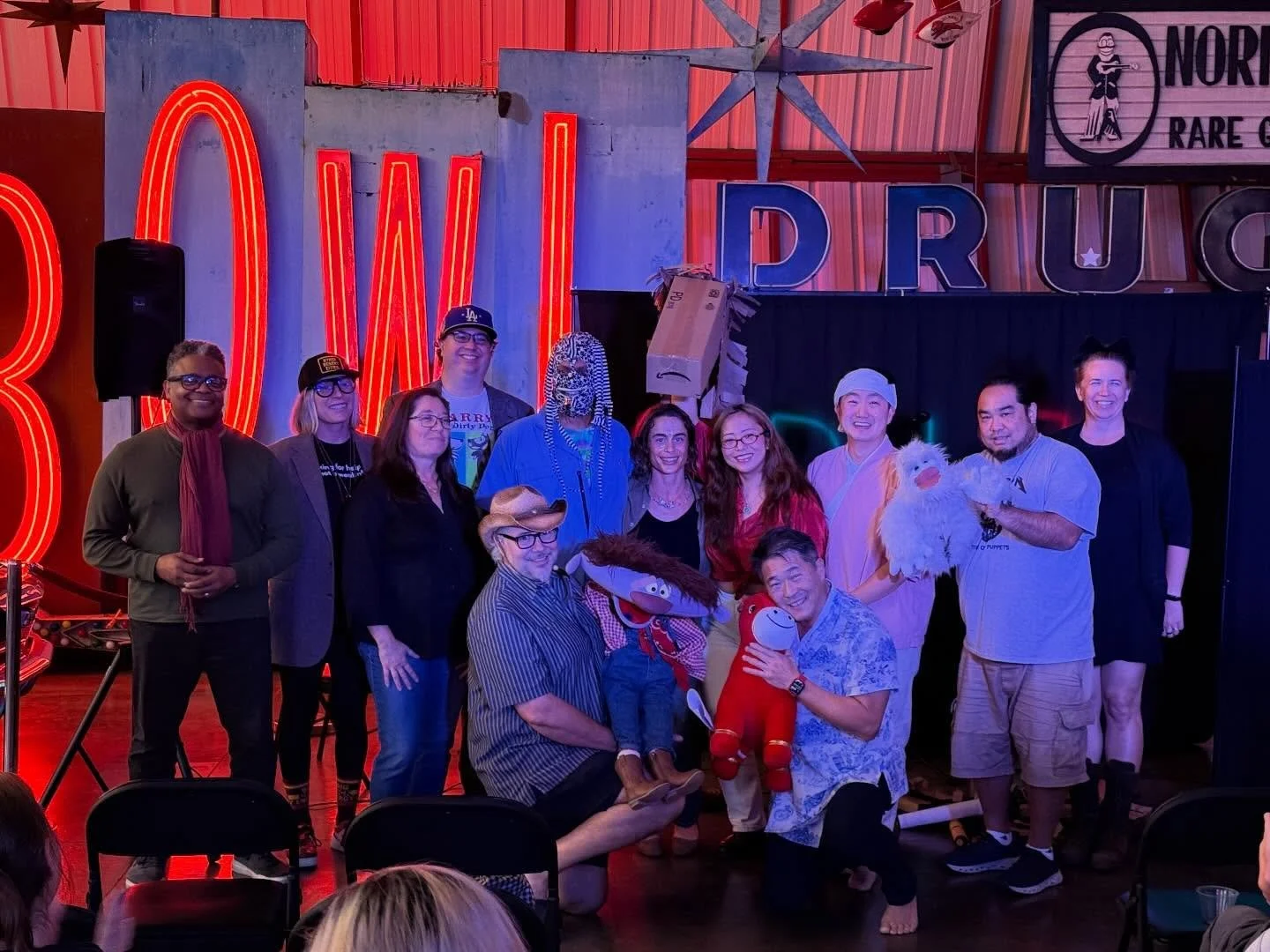 Group of people posing together at a puppetry or entertainment venue with colorful neon signs and puppets in the background.