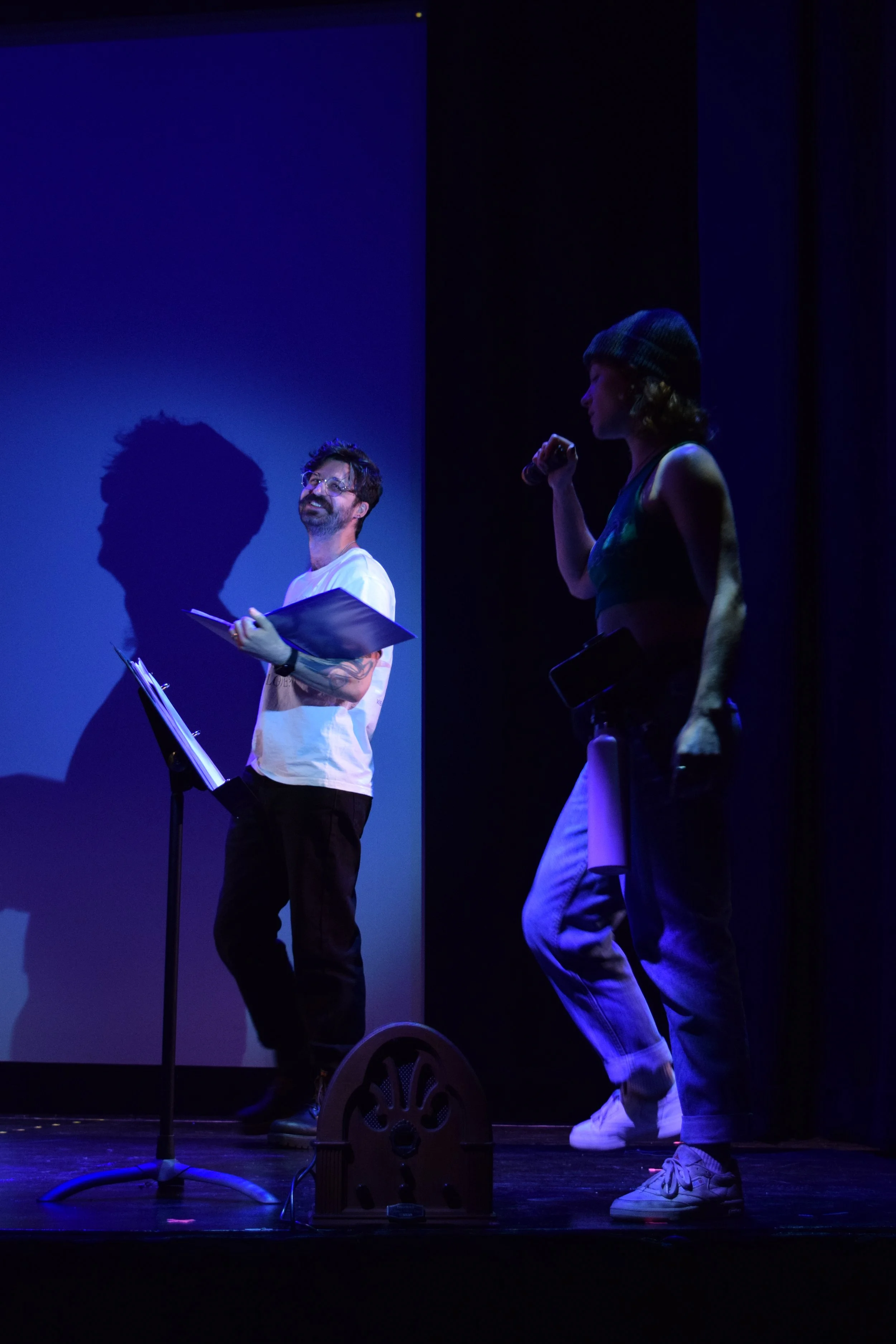 A man with glasses and a beard holding a script or music stand on stage, facing a woman with a beanie and a microphone, with stage lighting creating shadows.