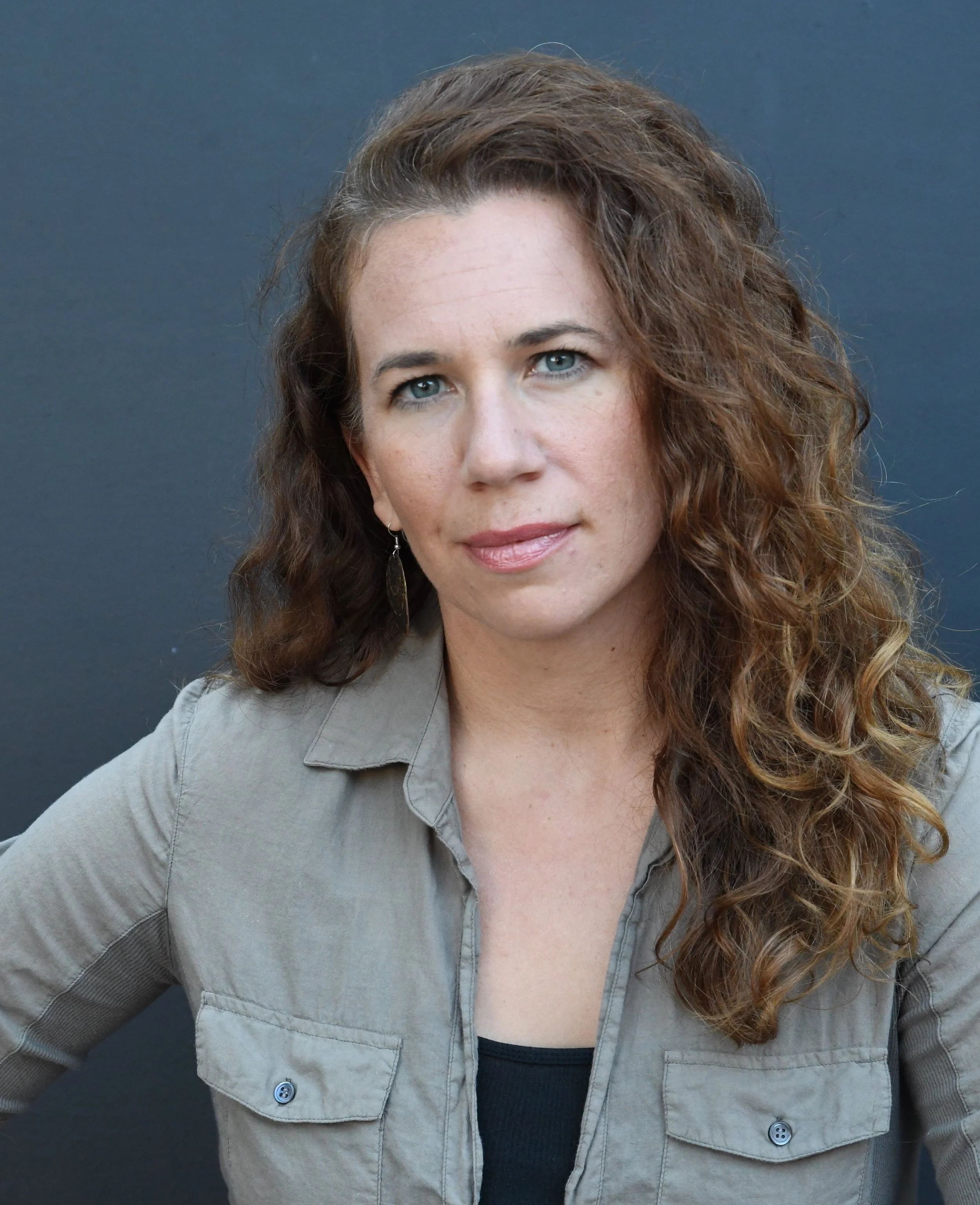 A woman with curly brown hair, blue eyes, and light skin, wearing a gray jacket over a black shirt, has a neutral expression, against a plain dark background.