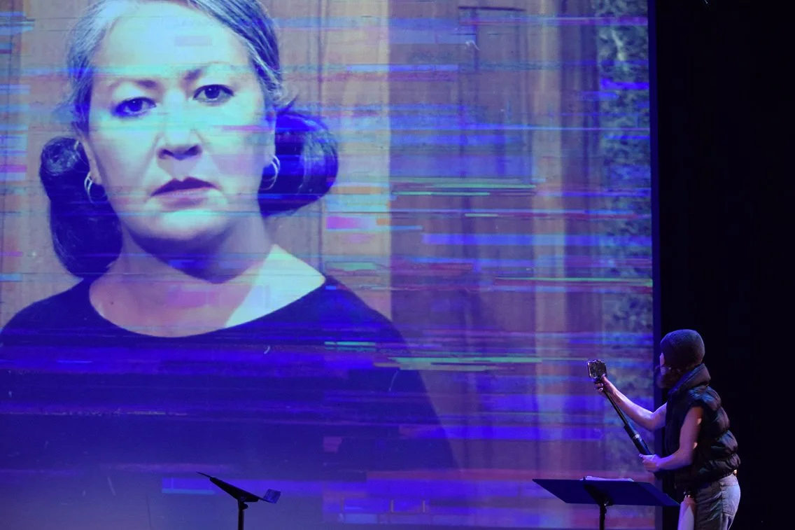 A person on stage using a selfie stick to take a photo of a large screen displaying a woman's face with blue glitches.