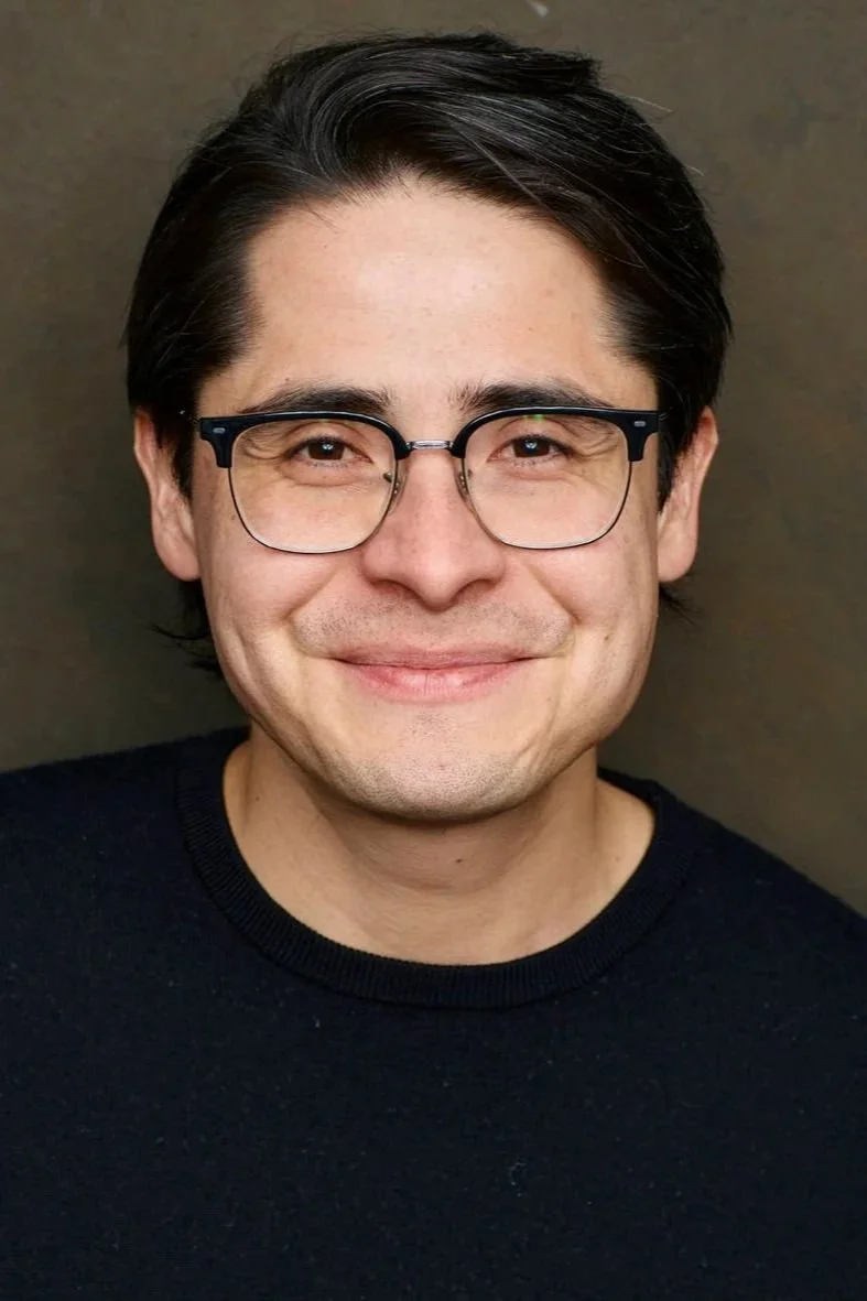 Close-up of a smiling man with dark hair, glasses, wearing a black shirt, against a plain background.
