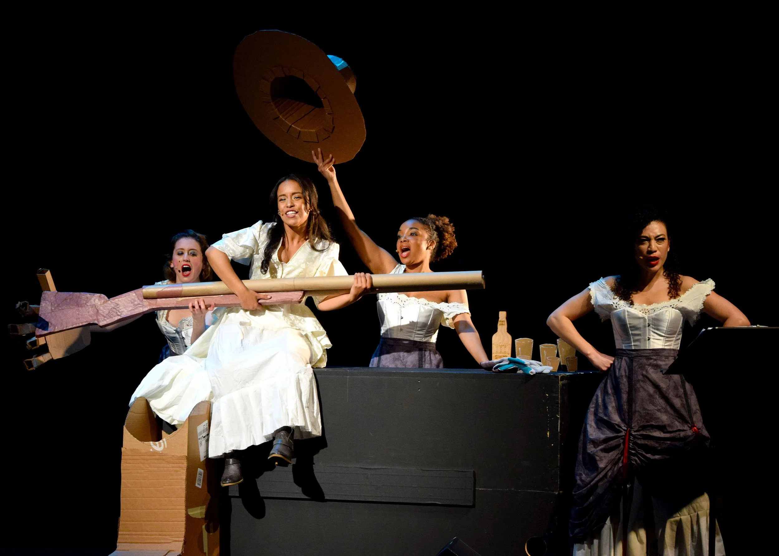 A theatrical performance with four women in costumes, one playing a stringed instrument, on a dark stage with props including a large pink umbrella, a cardboard box, a wooden stick, and glasses.