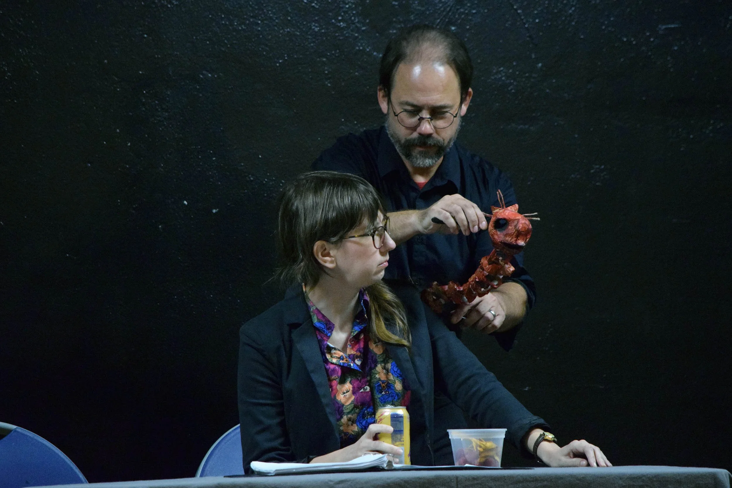 Two individuals sitting at a table; one holding a drink and the other holding a small-model red dinosaur.