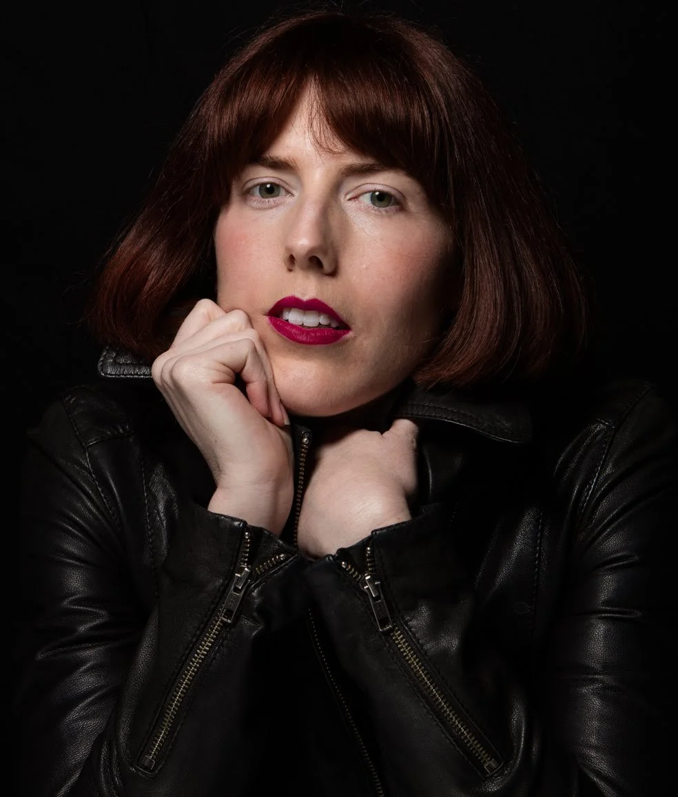 A woman with shoulder-length red hair, wearing a black leather jacket, looks directly at the camera with her chin resting on her hands, against a dark background.