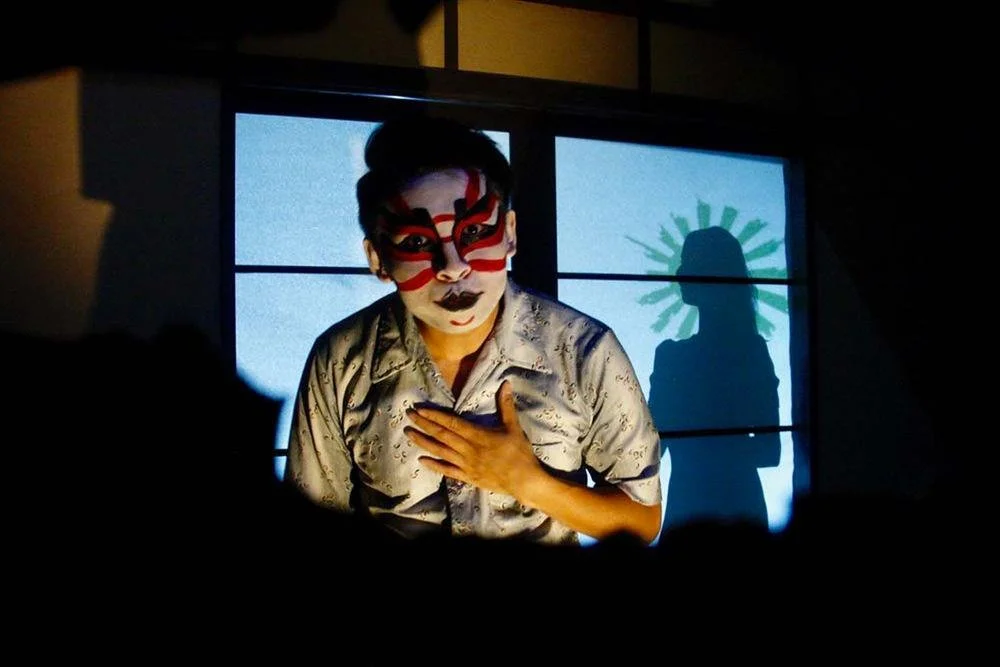 Performer with face paint and costume on stage, hand on chest, silhouette with halo backdrop projected behind