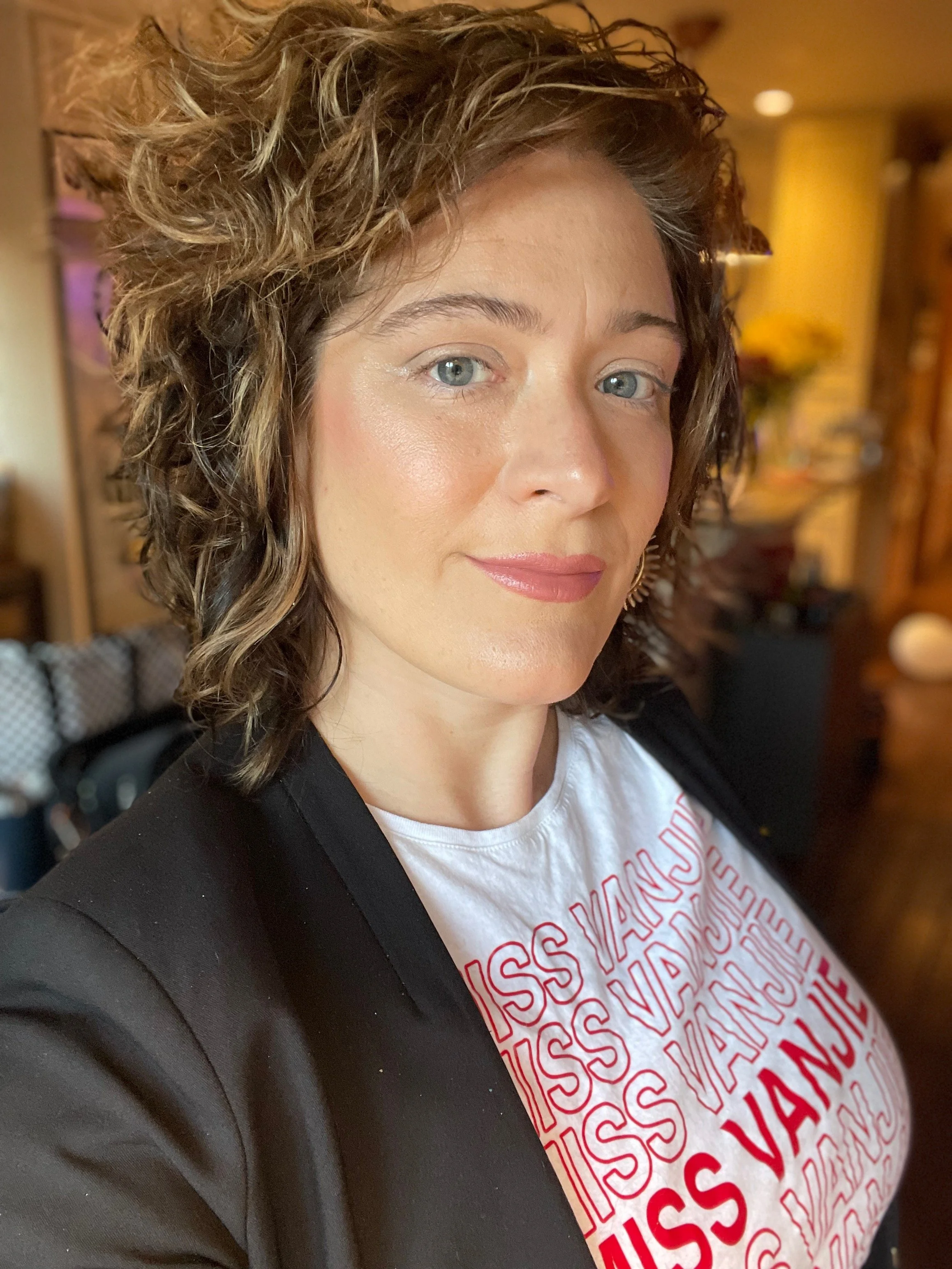 A woman with curly brown hair and light-colored eyes, wearing a white T-shirt with red text and a black jacket, standing indoors with a warm yellow-brown background.