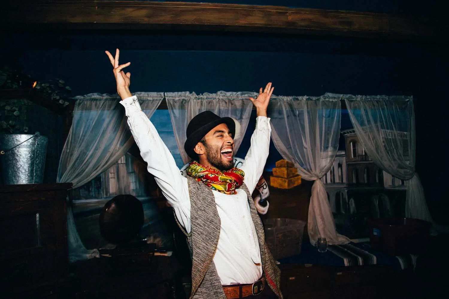 A happy man wearing a black hat, white shirt, and colorful scarf, raising his arms in celebration or excitement indoors with wooden walls and white curtains in the background.