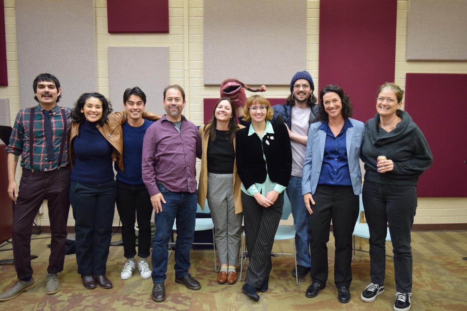 Group of nine diverse people standing together in a room, smiling, with a puppet resembling a red creature with a long nose in the background.