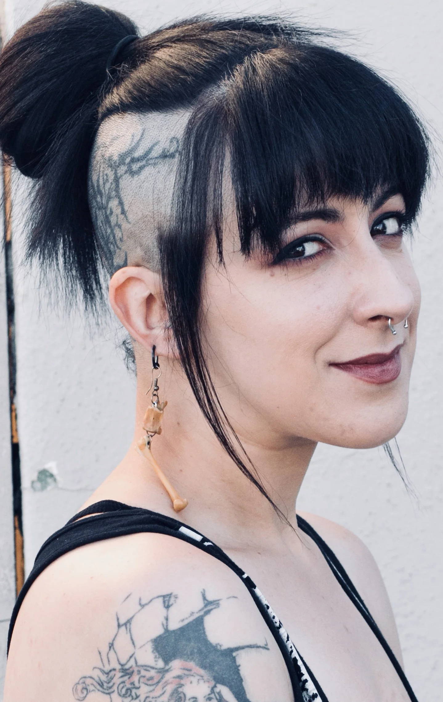 A person with black hair styled to the side, including a shaved section with a tattoo of a tree on the scalp. They have multiple facial piercings, earrings, and a tattoo on the arm. They are wearing a black and white striped top and an orange headphone around the neck, standing against a plain background.