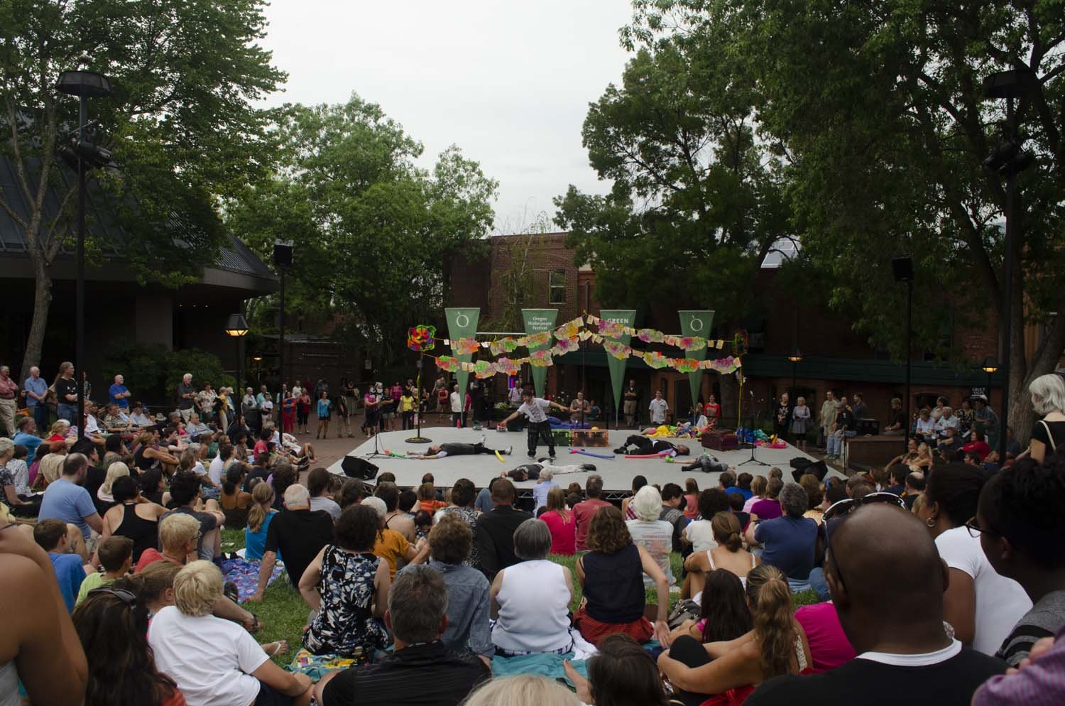 Crowd gathered around an outdoor stage with performers and colorful decorations in a park setting.