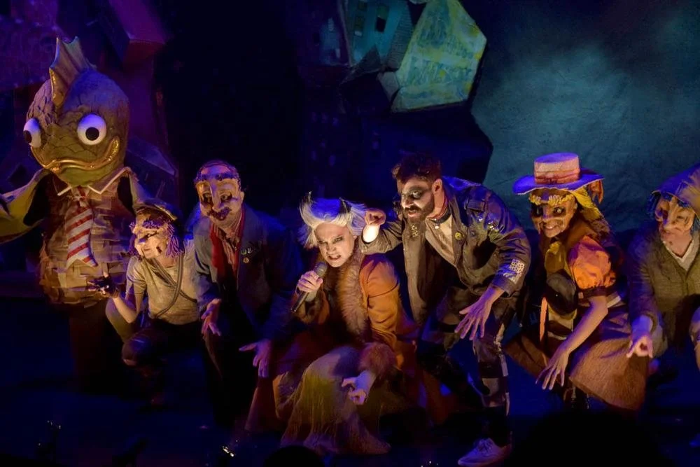 Theatrical stage with actors in costumes, including a character in a fish costume and actors wearing masks, with dramatic lighting and colorful background.